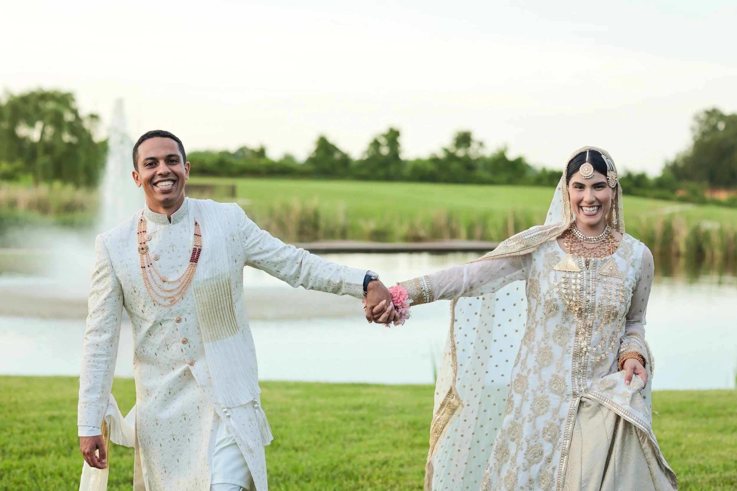 A happy bride and groom in traditional Indian wedding attire outdoors near a pond with a fountain and green landscape in the background, holding hands and smiling.