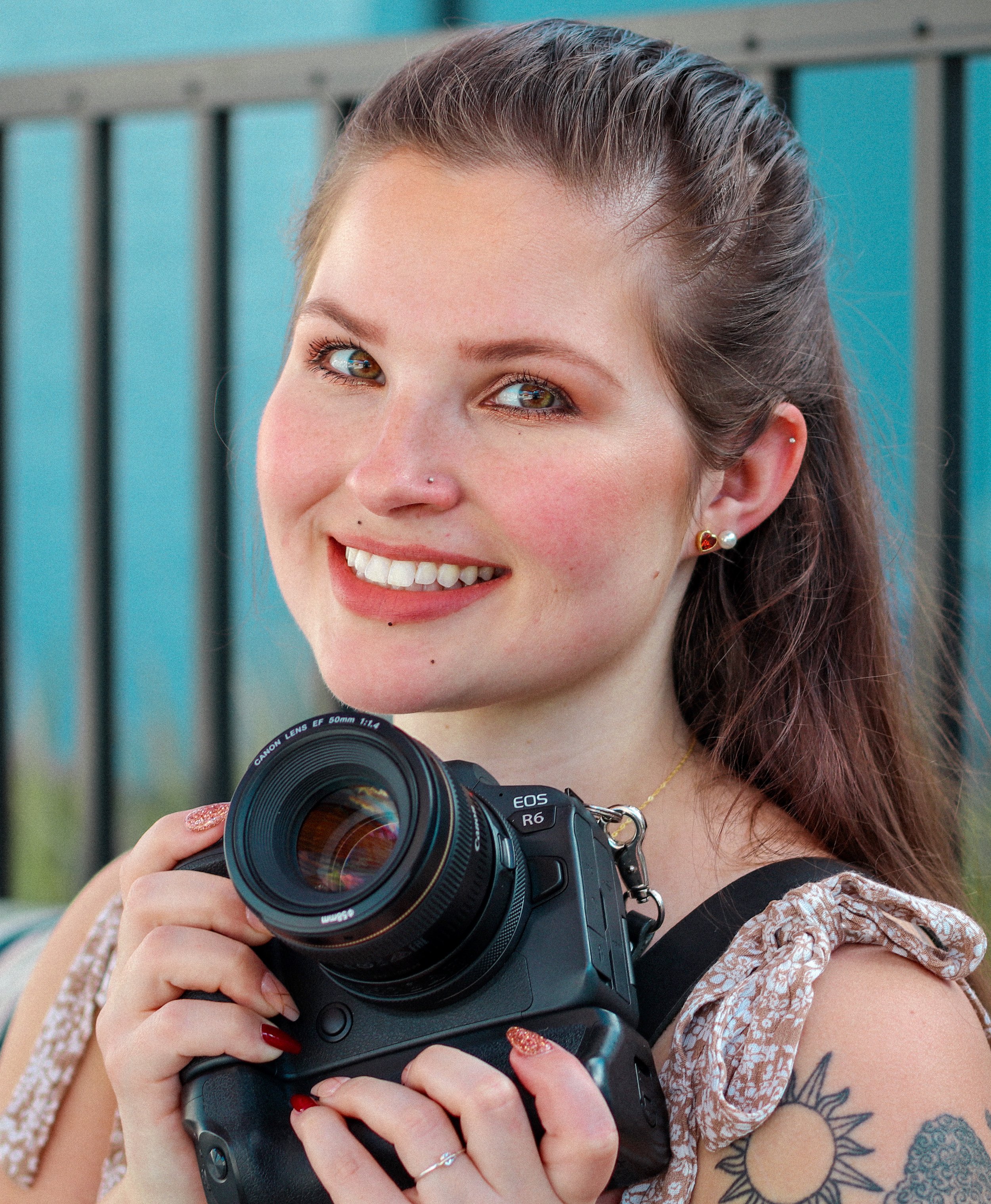 A smiling woman holds a professional camera with a large lens, standing outdoors near a blue building or fence.