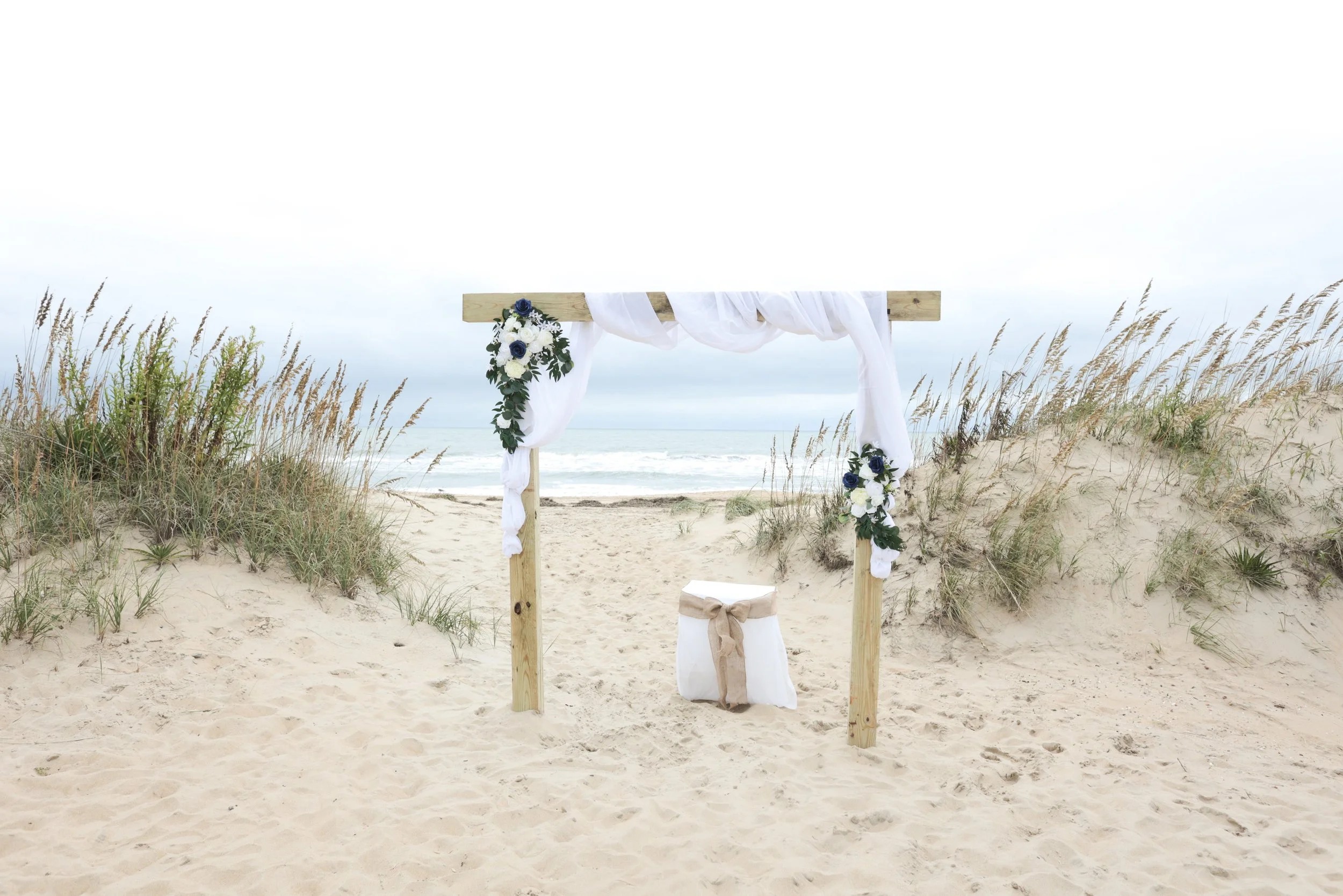 Beach wedding arch decorated with white fabric and navy and white flowers, with a small table draped with white cloth and tied with a beige bow, on sandy dunes with grass, overlooking the ocean under a cloudy sky.