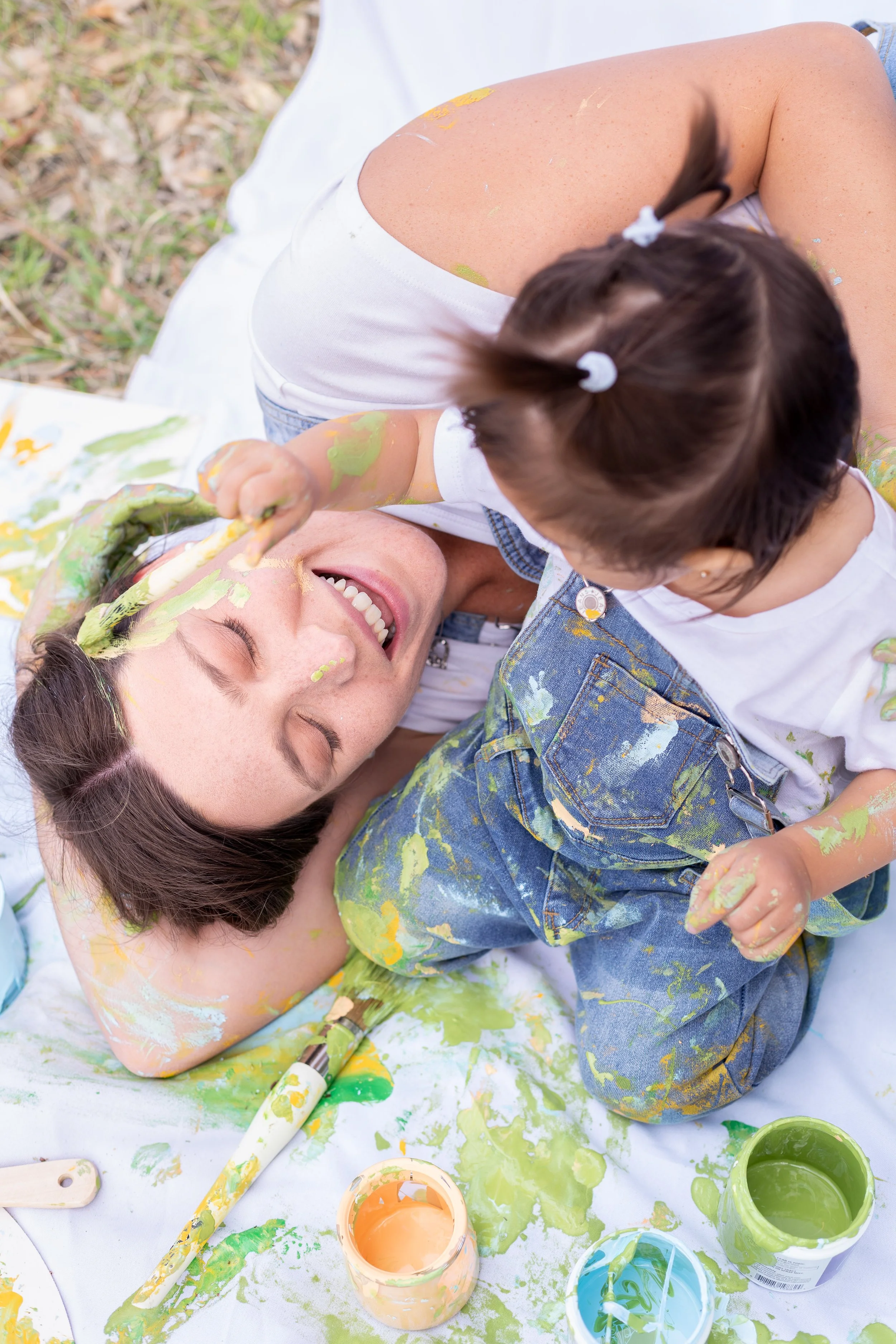 A woman lying on the ground with closed eyes, smiling, being playfully covered in green and yellow paint by a young child with brown hair tied in pigtails. The child is also covered in paint and holding a paintbrush. There are open jars of green, yellow, and blue paint and a paintbrush on the white cloth-covered ground.