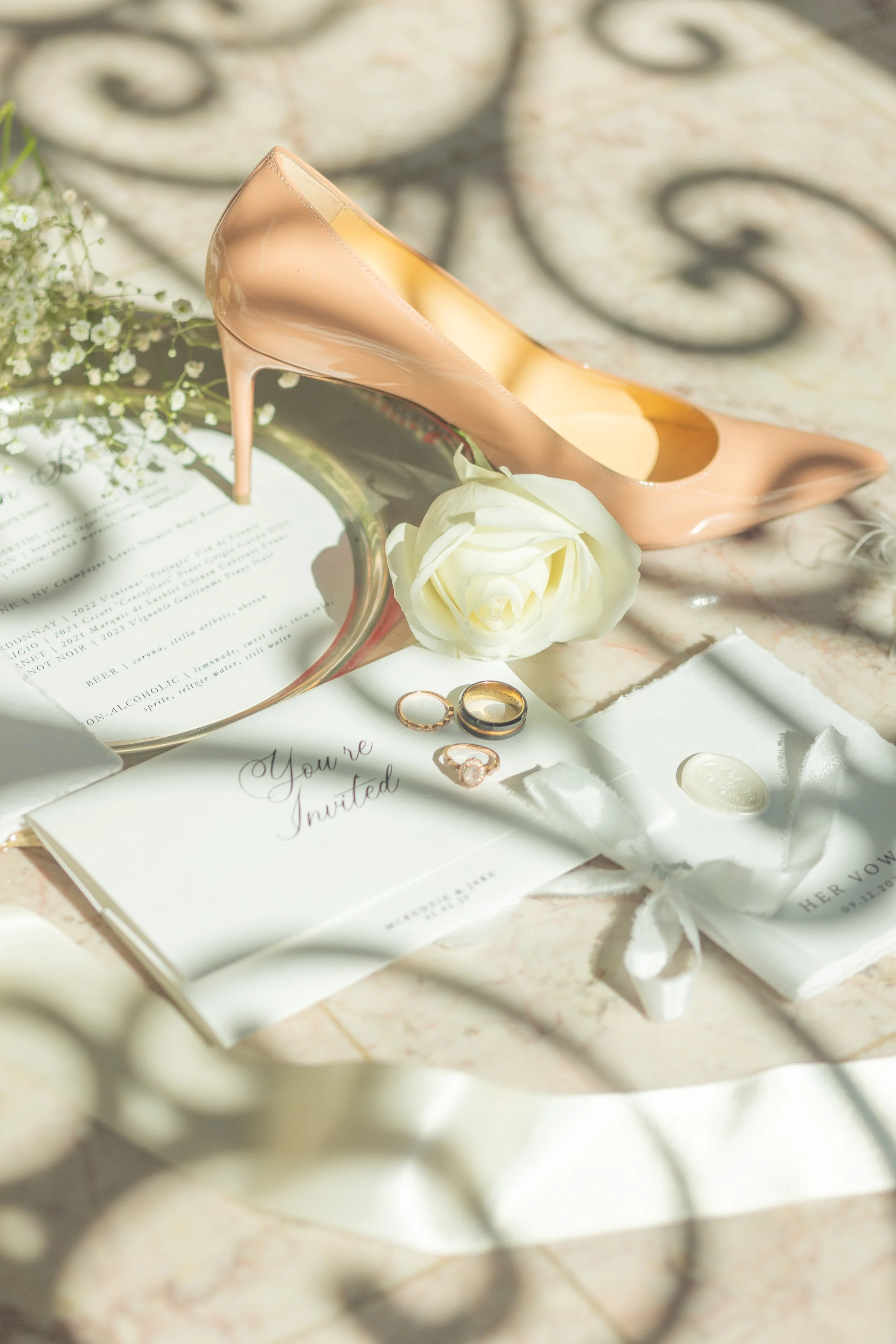 Elegant bridal flat lay featuring a blush stiletto, white rose, gold wedding bands, wax-sealed vow booklet, and wedding invitation. Light and airy detail photo ideal for luxury wedding inspiration and editorial styling.