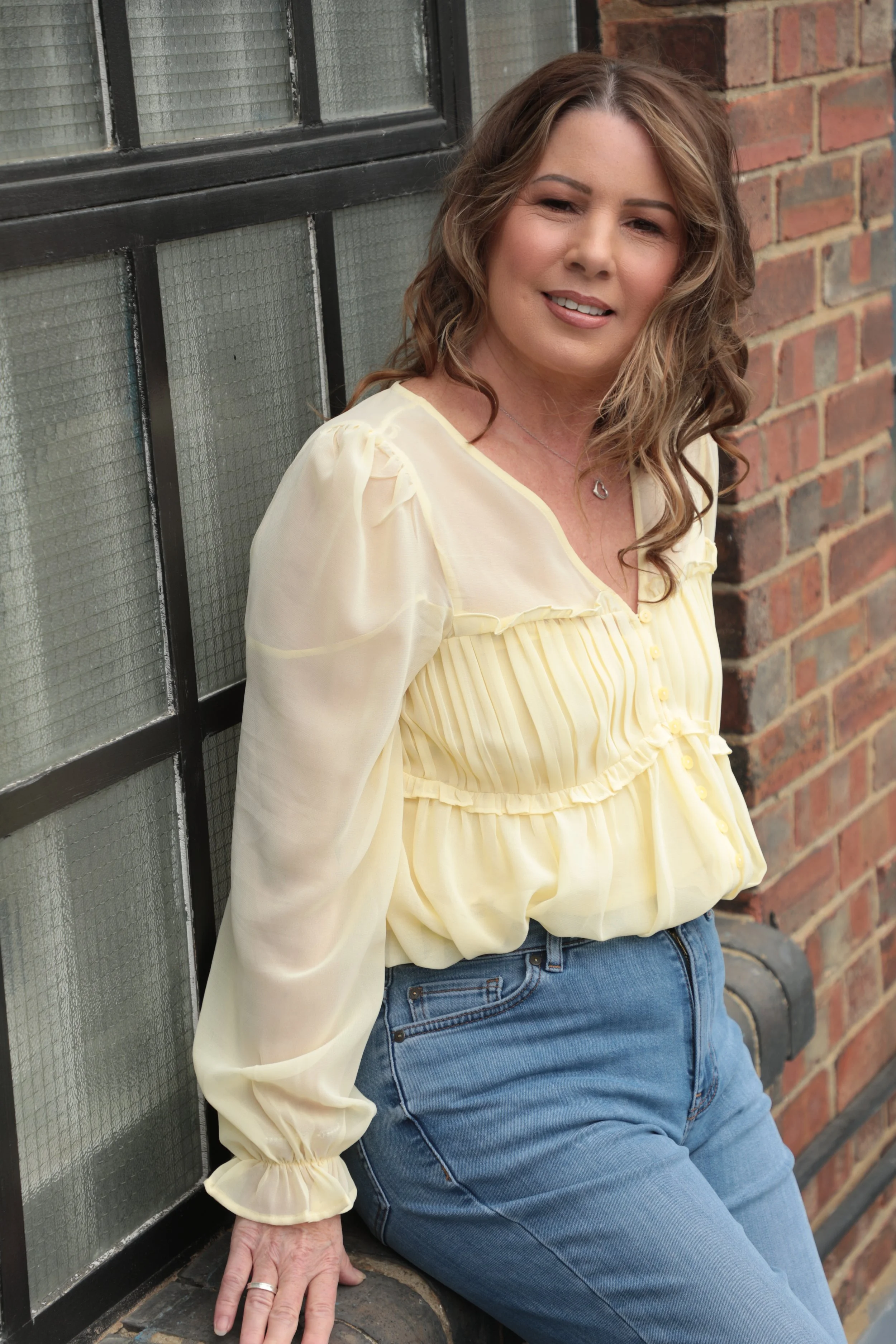 A woman with wavy brown hair wearing a yellow blouse and blue jeans is sitting on a brick sidewalk against a brick wall and black-framed glass window.