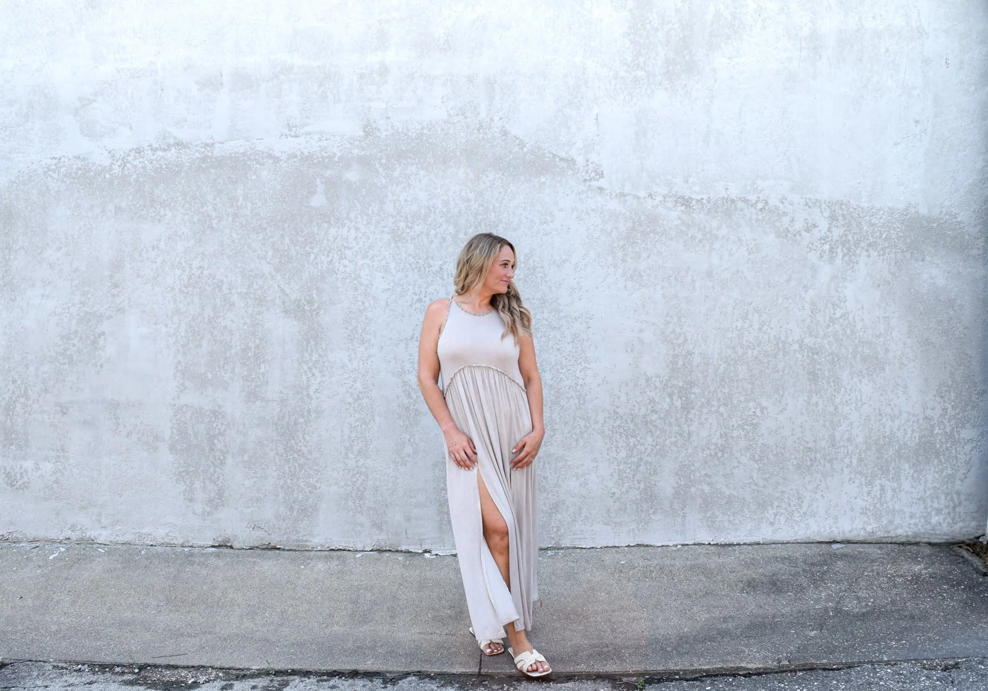 A woman in a beige dress with a slit standing against a textured white wall.