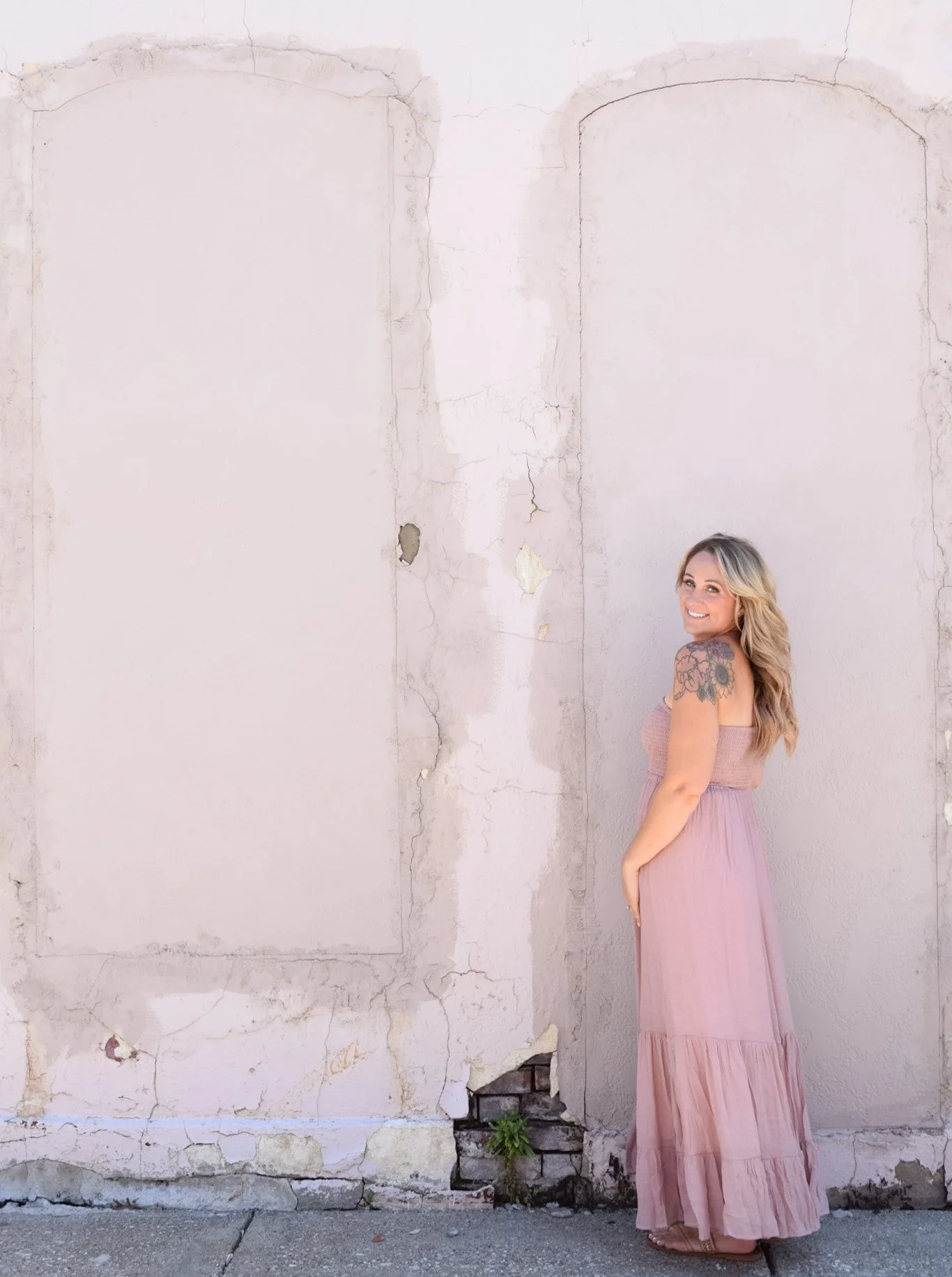 A woman with long blonde hair in a strapless pink dress standing against a weathered, off-white wall with peeling paint and exposed bricks near the bottom, smiling and looking at the camera.