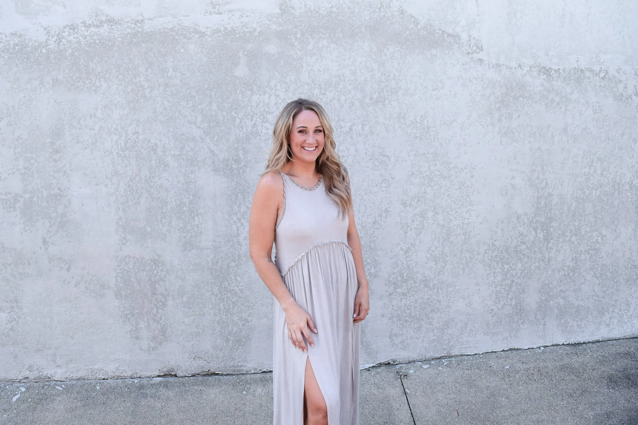 A smiling woman with blond hair in a sleeveless, light-colored gown standing against a plain, textured concrete wall.