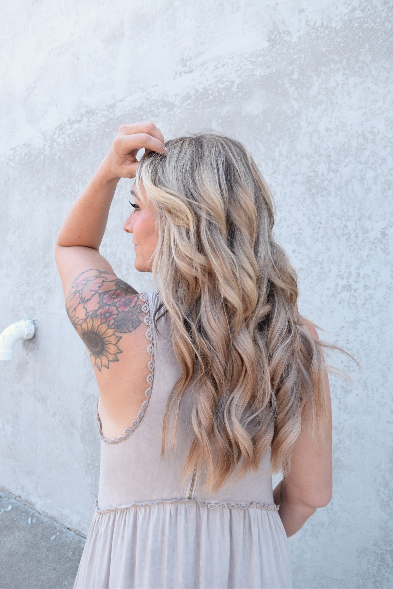 Side view of a woman with long wavy blonde hair, wearing a sleeveless beige dress, standing against a textured light wall with her left arm raised, touching her hair, revealing a tattoo of sunflowers and flowers on her upper arm.