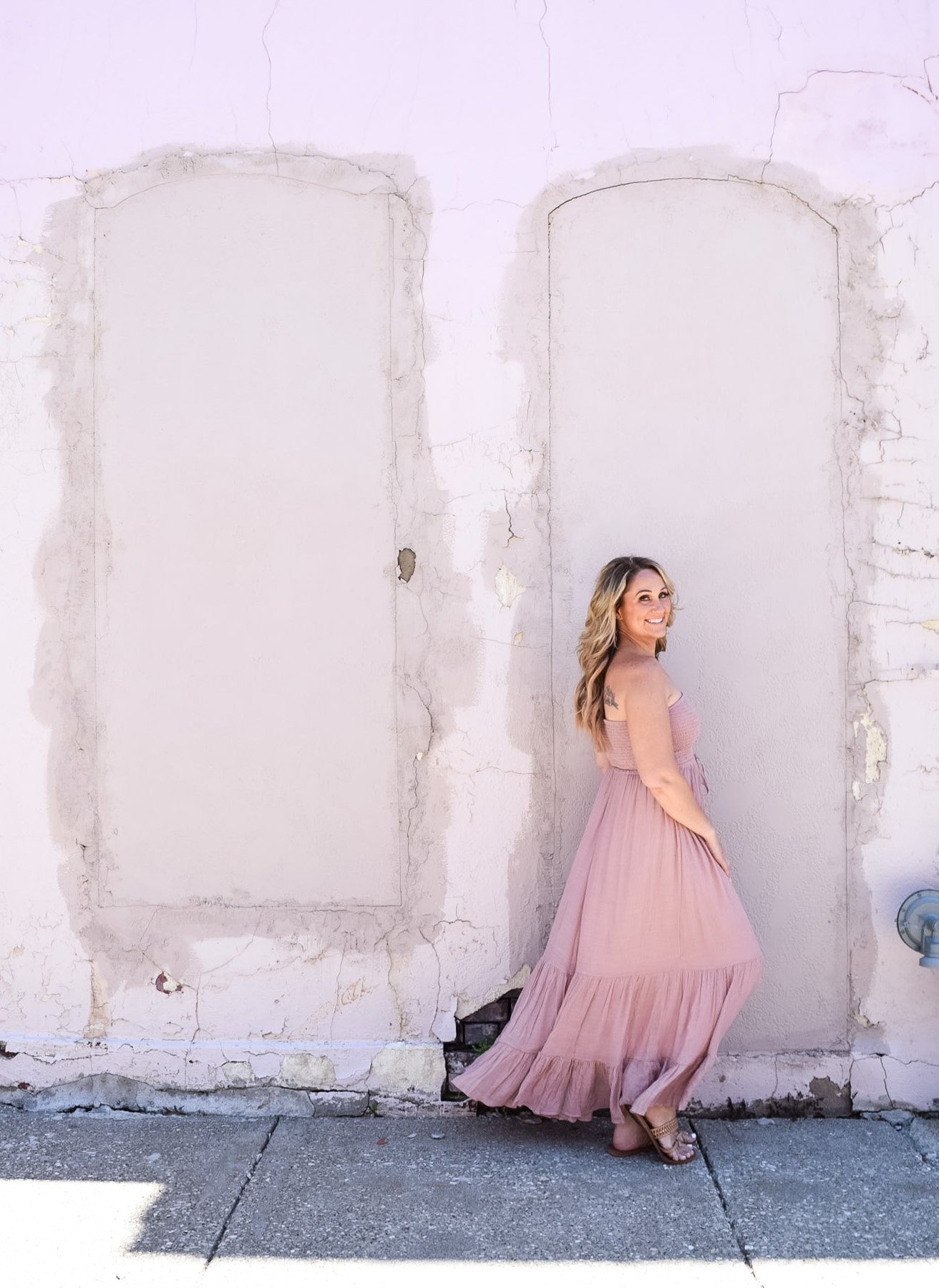 A woman in a pink maxi dress standing against a weathered, pale pink wall with empty rectangular wall outlines, smiling and looking back.