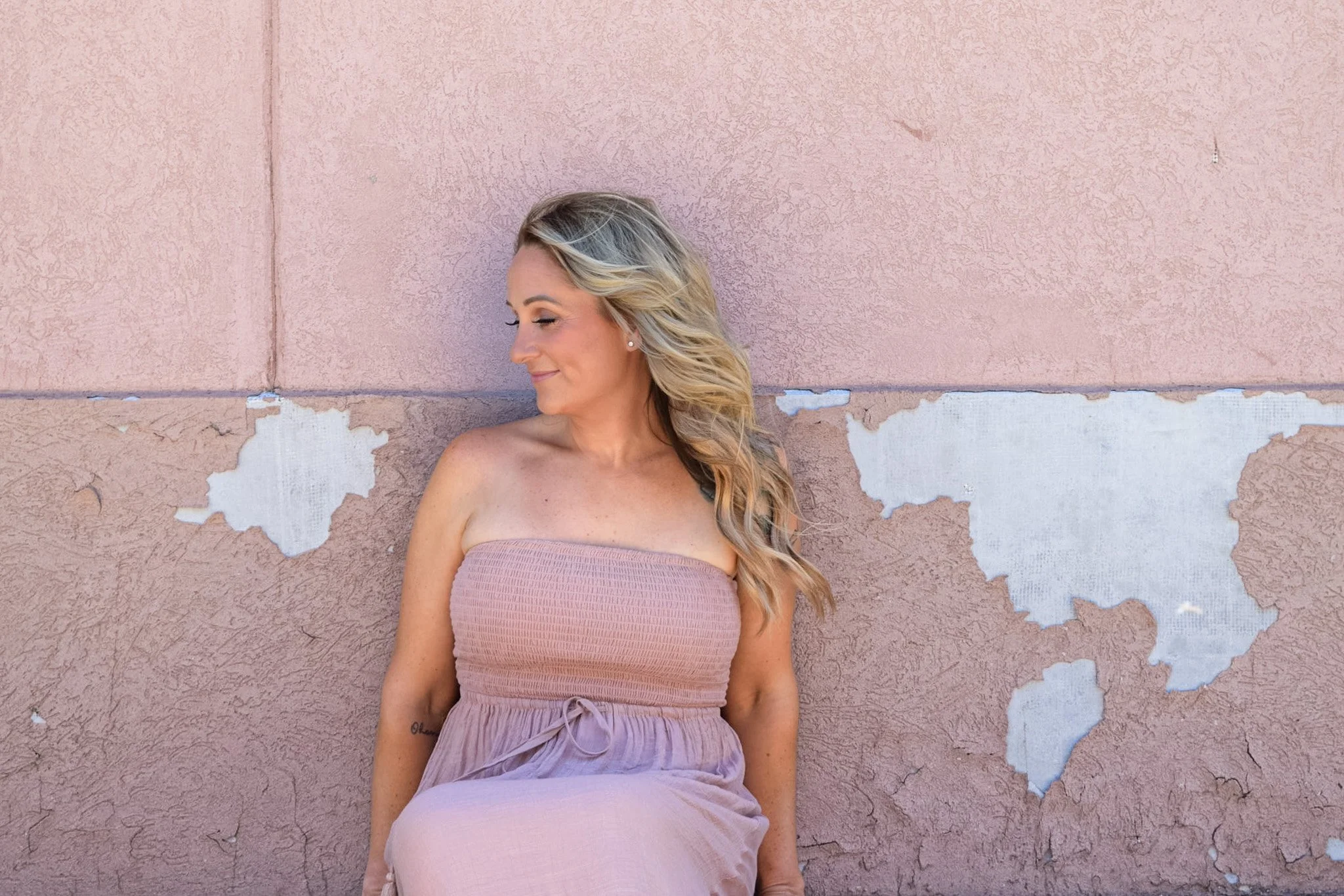 Woman with wavy blonde hair in a strapless pink dress leaning against a pink wall with peeling paint.