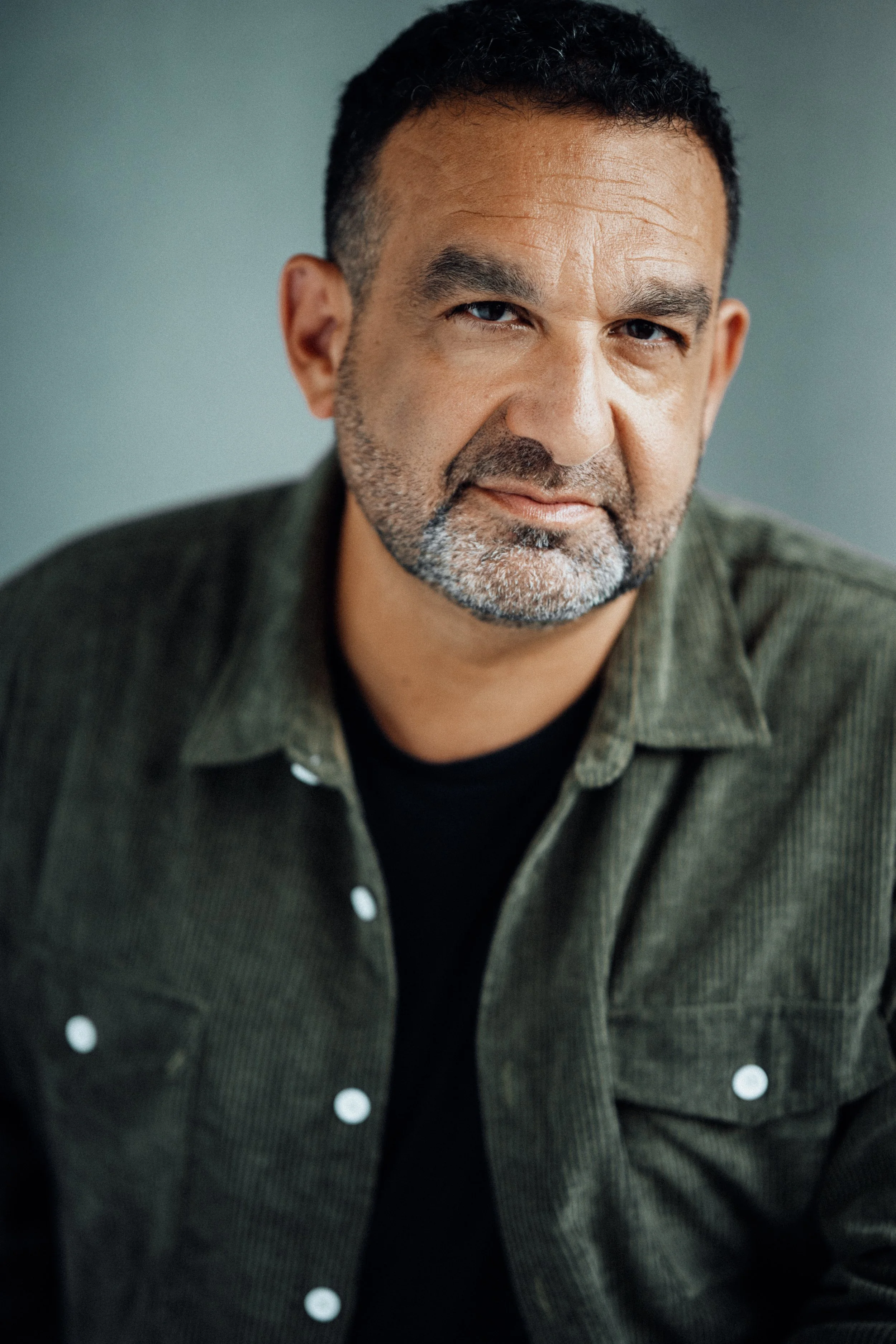 Close-up of a middle-aged man with short dark hair, a beard with some gray, wearing a dark green corduroy shirt over a black shirt, looking confidently at the camera.