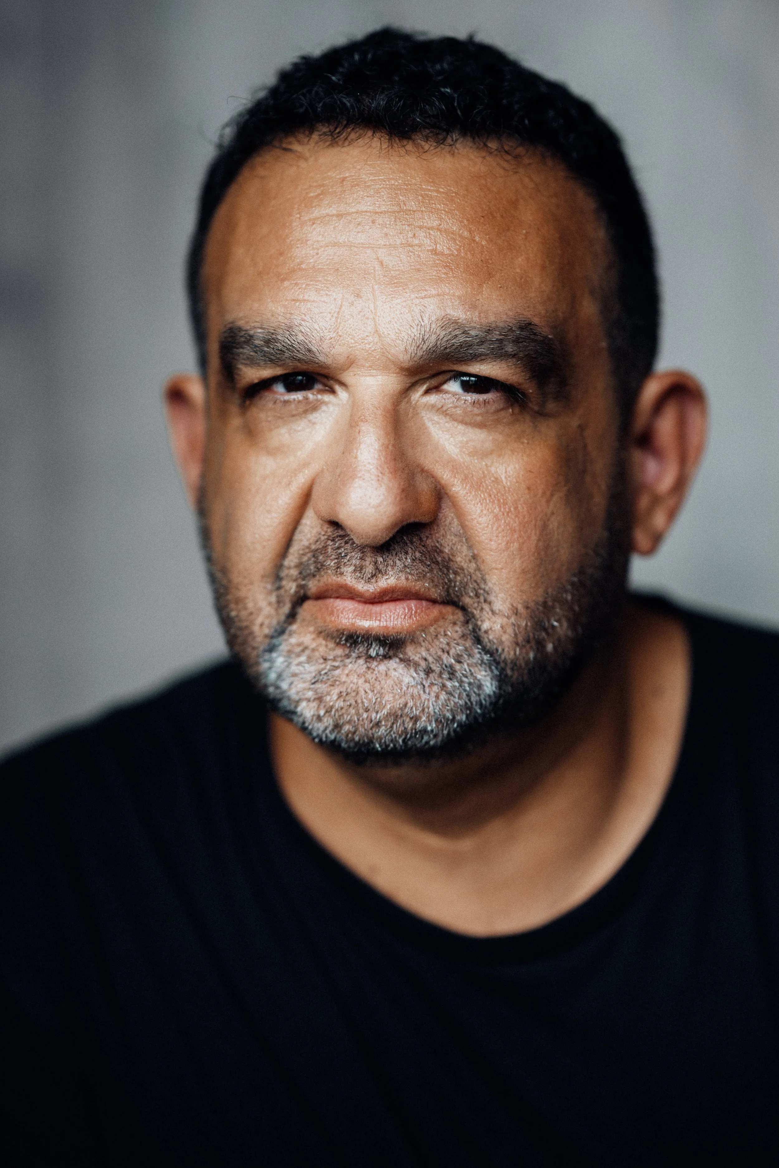 Close-up of a mature man with a beard, short dark hair, wearing a black shirt, looking directly at the camera with a serious expression.