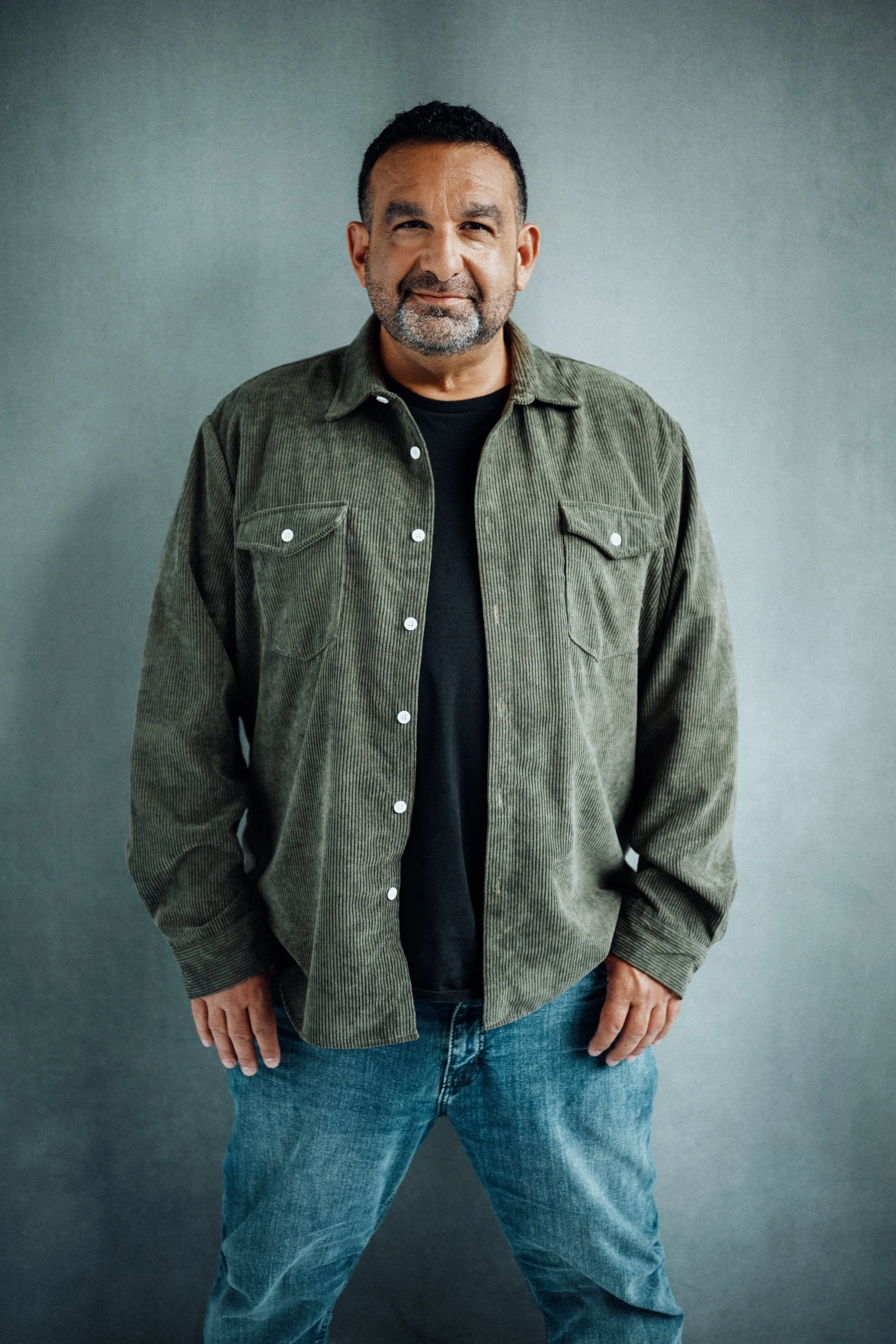 A middle-aged man with short dark hair, a beard, and a friendly smile, standing against a plain gray background. He is wearing a green corduroy button-up shirt over a black t-shirt and blue jeans.