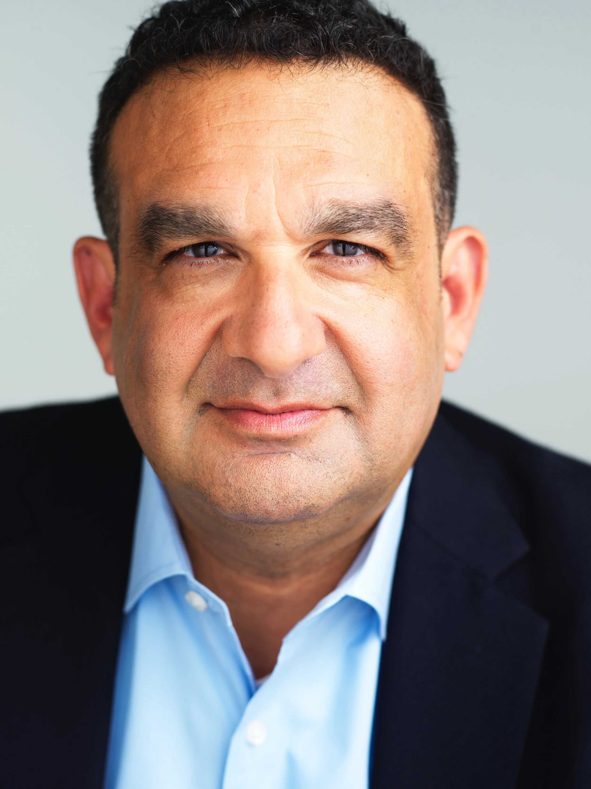 Close-up of a middle-aged man with short, dark hair, wearing a light blue collared shirt and a dark suit jacket, looking directly at the camera with a neutral expression.