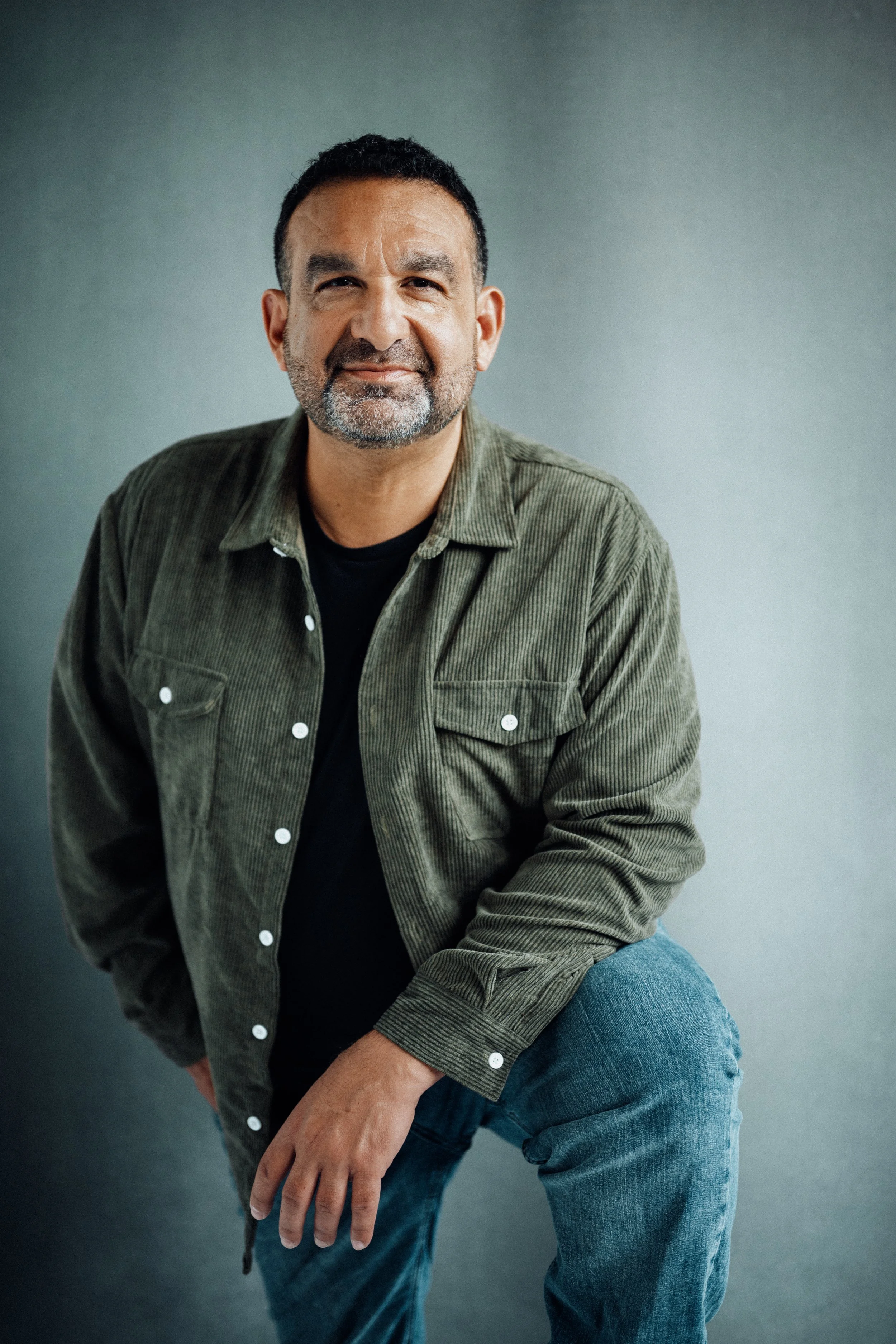 A middle-aged man with dark hair, a beard, and a friendly smile, dressed in a green corduroy jacket over a black t-shirt and blue jeans, posing casually against a plain gray background.