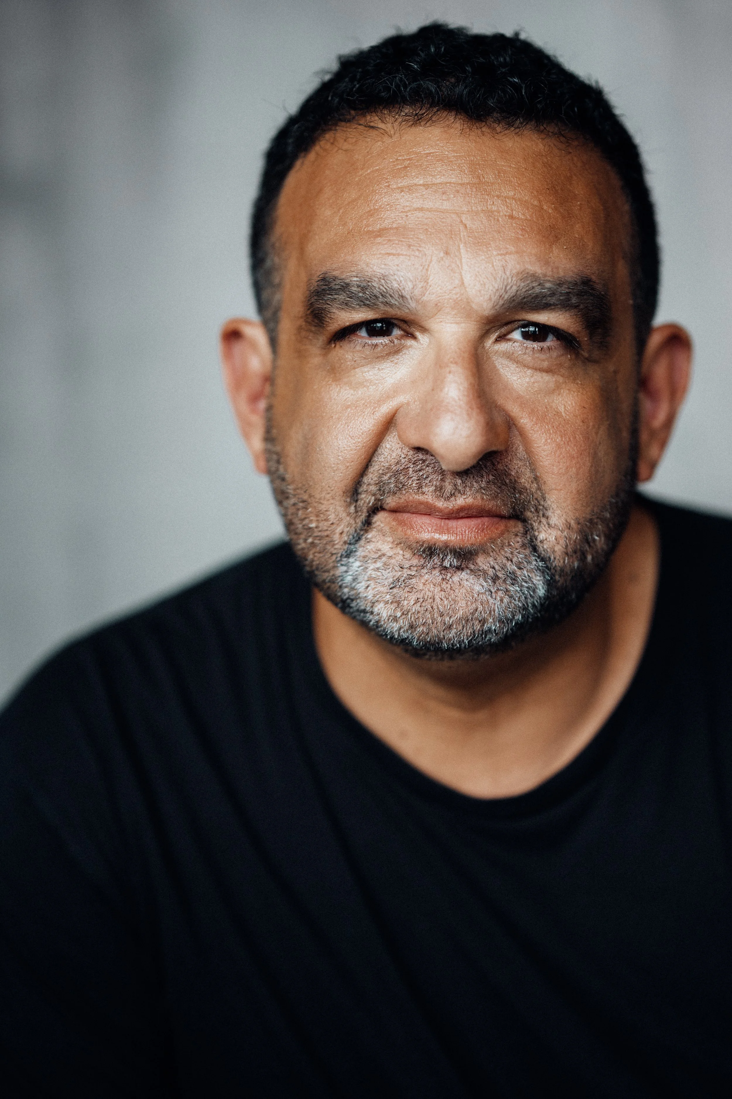 Close-up headshot of a middle-aged man with short, dark hair, brown eyes, and a salt-and-pepper beard, wearing a black shirt, against a neutral background.