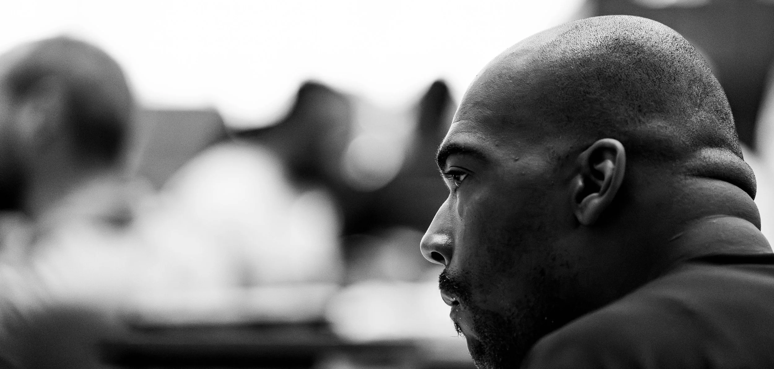Side profile of a young man with a shaved head, looking intently to the right, with blurred figures in the background, black and white photo.