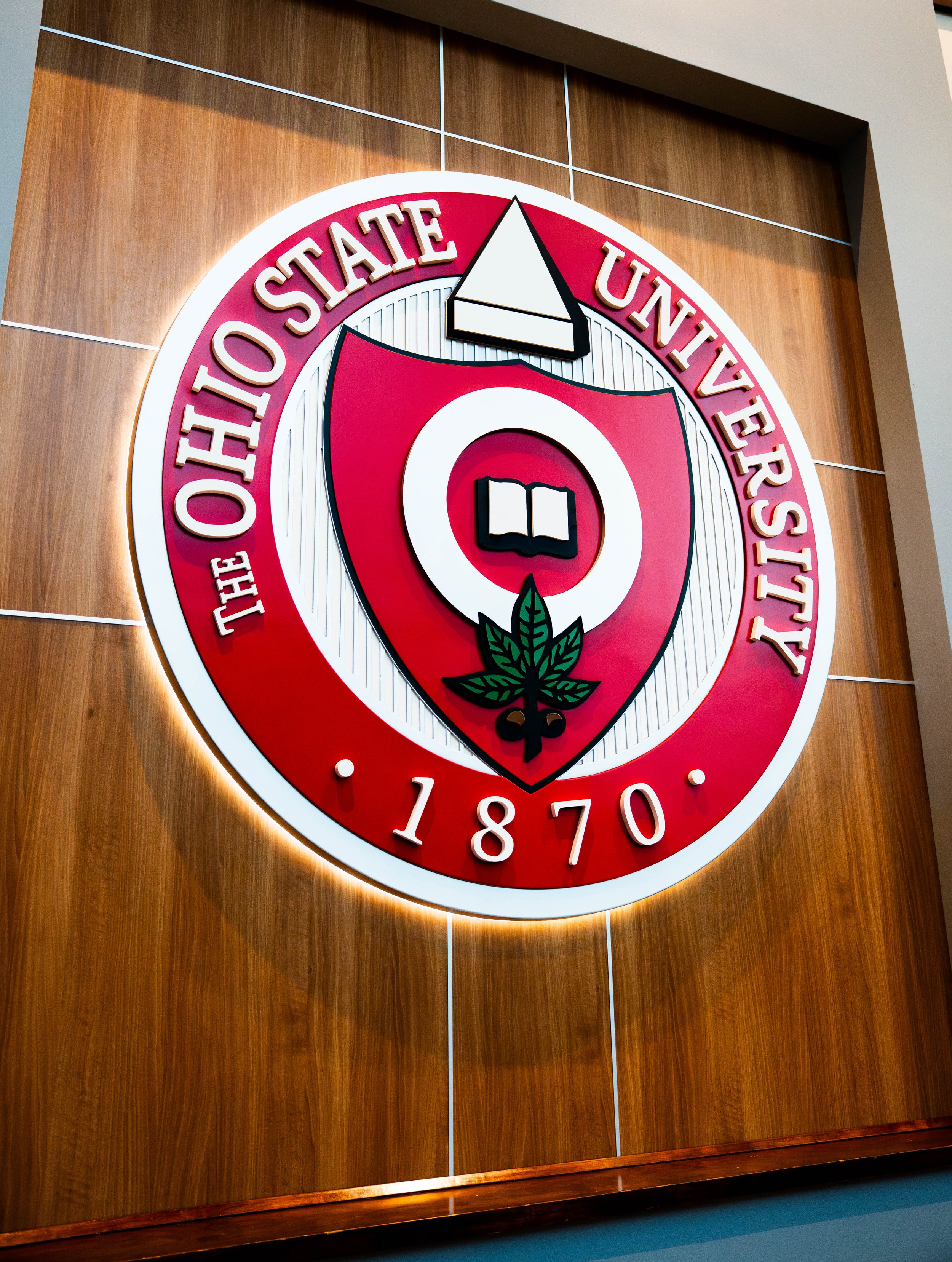 The Oklahoma State University seal on a wooden background, featuring a shield with a book and a maple leaf, with the year 1870 written below.