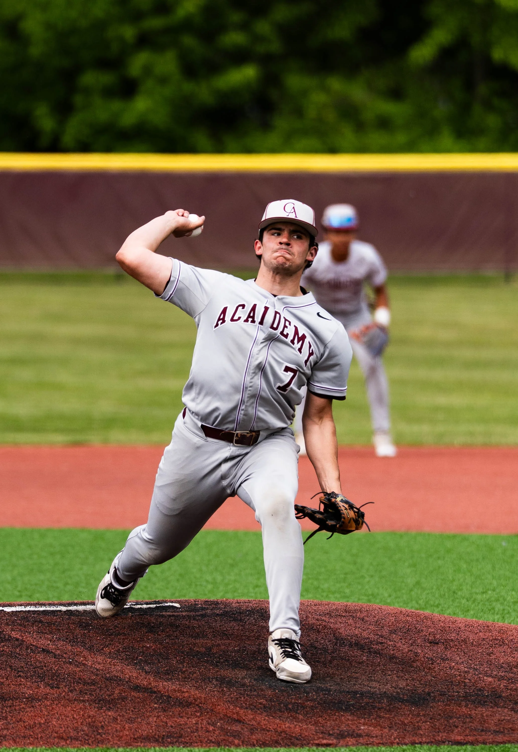 A baseball player is throwing a pitch on the mound during a game, with another player running in the background. The pitcher is wearing a gray uniform with 'ACADEMY' written across the chest, a baseball glove in his left hand, and a cap with the lett