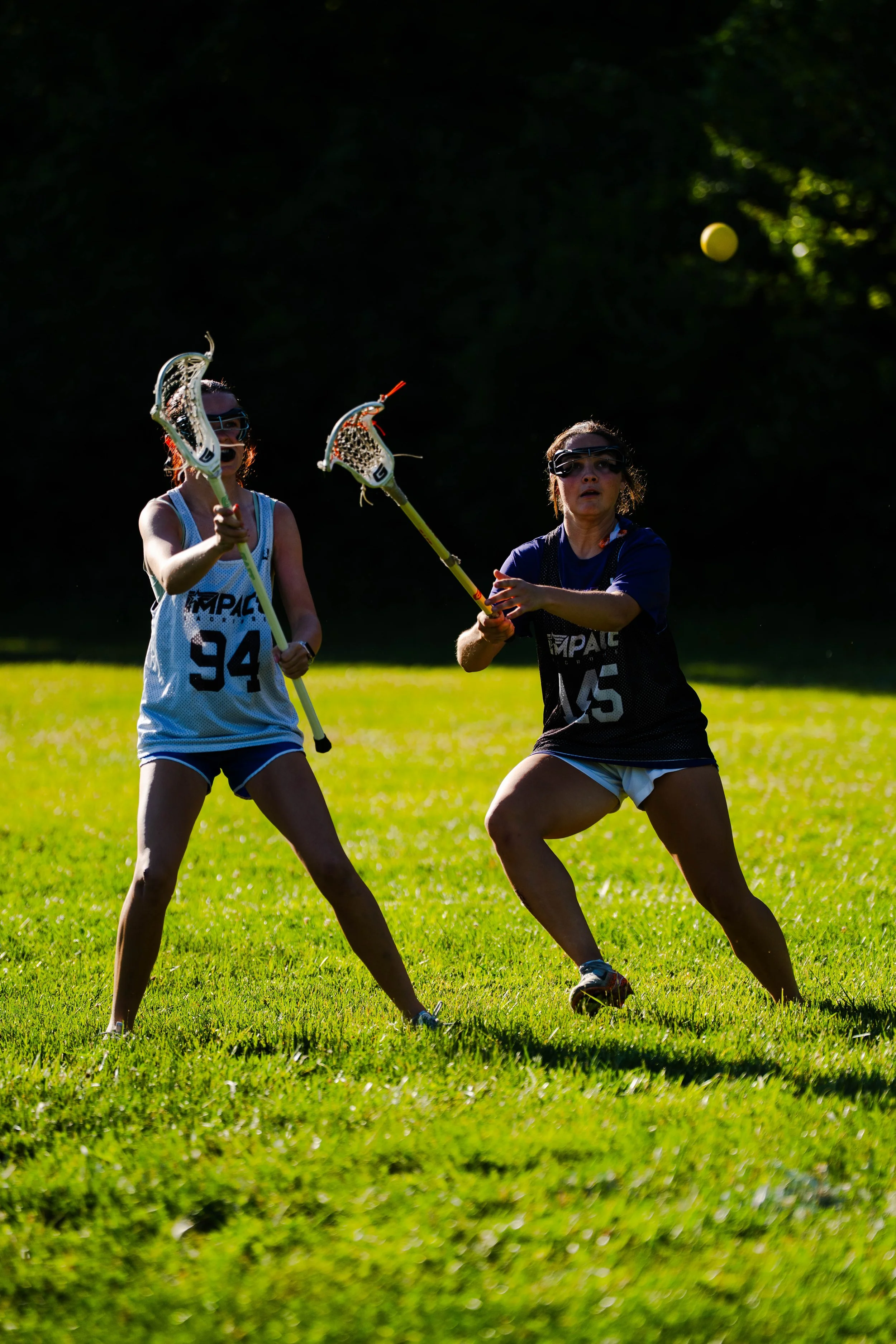 Two women playing lacrosse outdoors on a grassy field, preparing to catch or throw a yellow lacrosse ball, with a background of trees and sunlight filtering through the foliage.