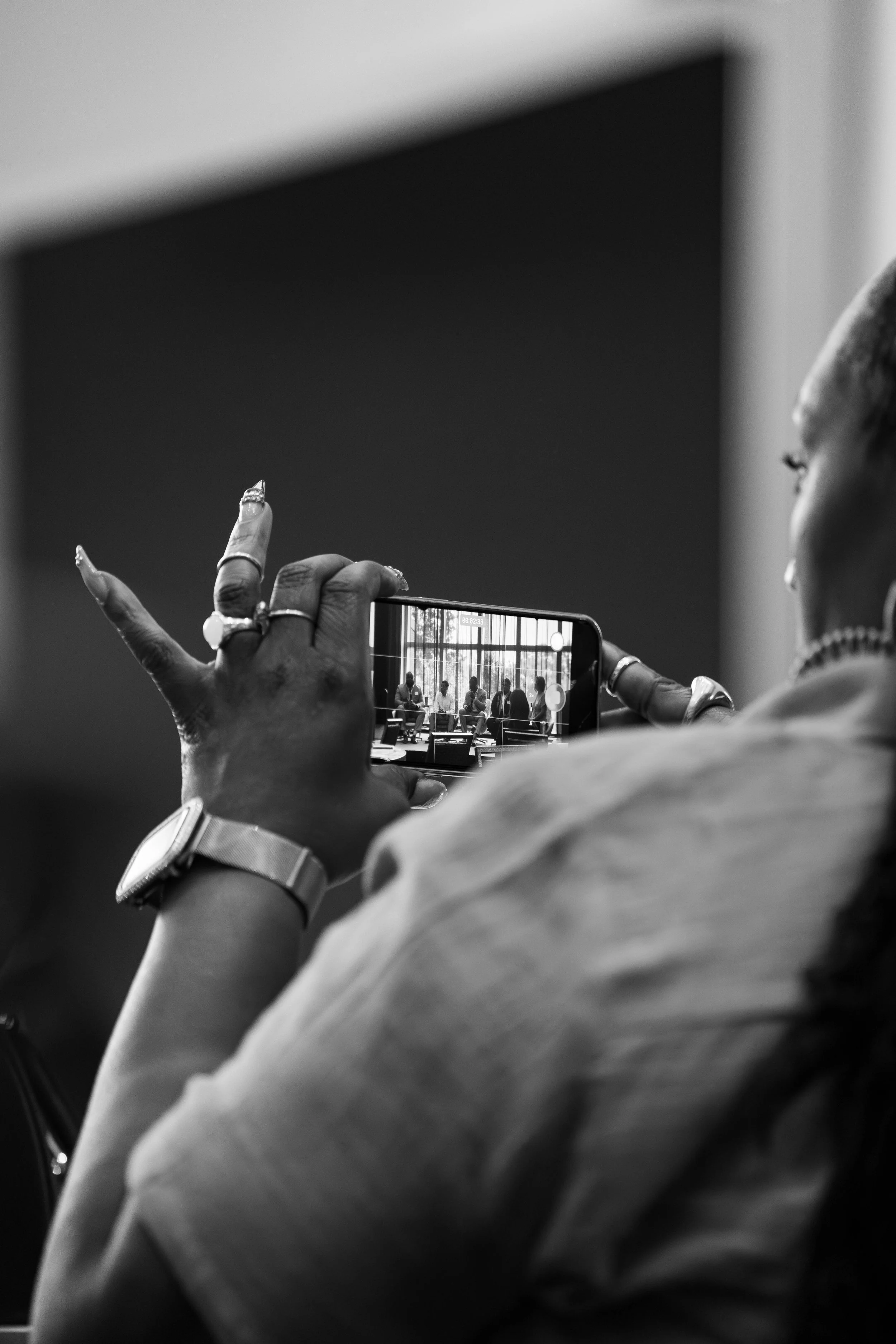 A close-up black-and-white photo of a person taking a picture with a smartphone, with the screen showing a group of people sitting at a long table in an indoor setting.