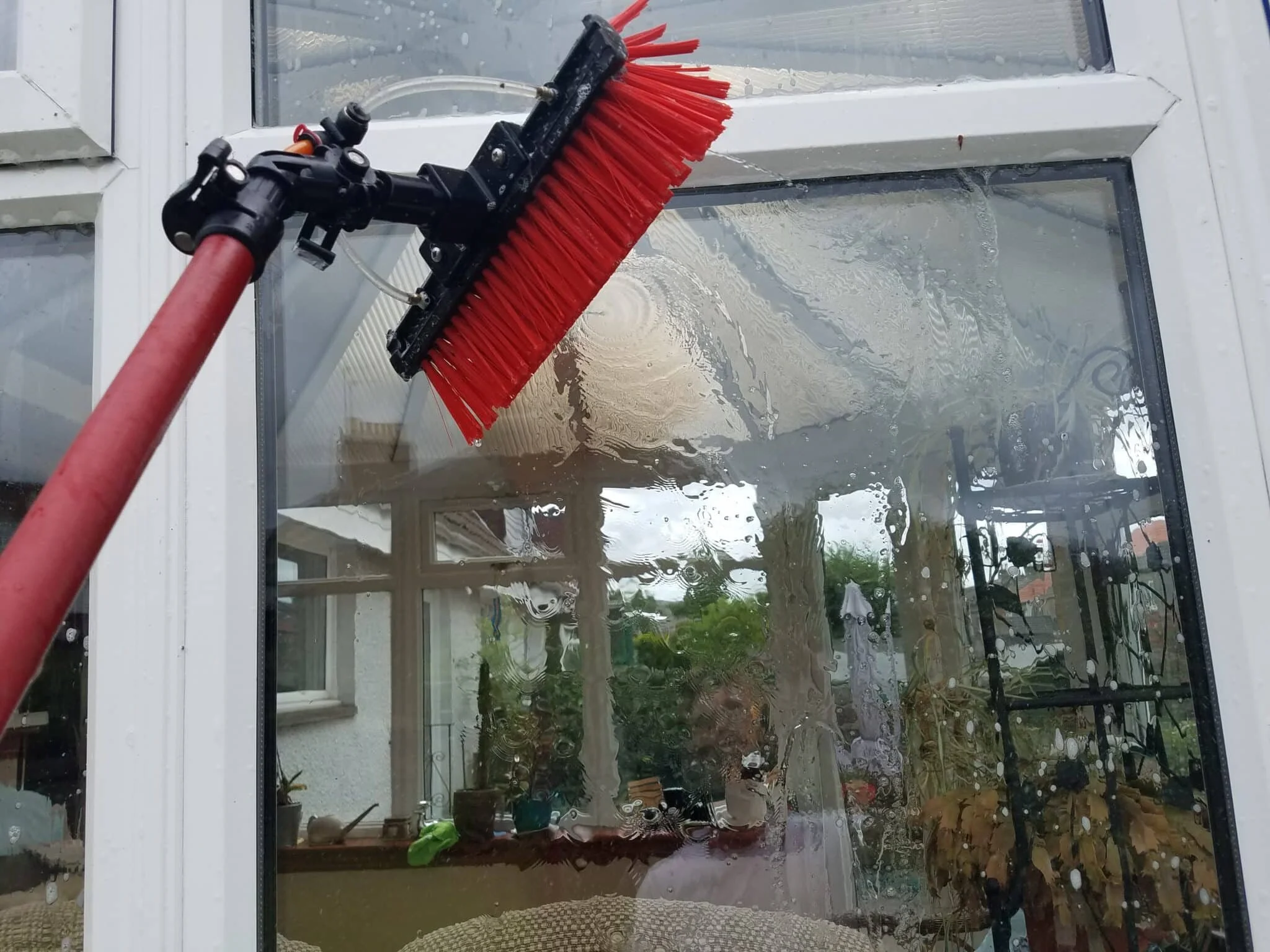 A window cleaning tool with red brushes attached to a handle, cleaning the glass of a large window in a residential area, with plants and outdoor furniture visible outside.