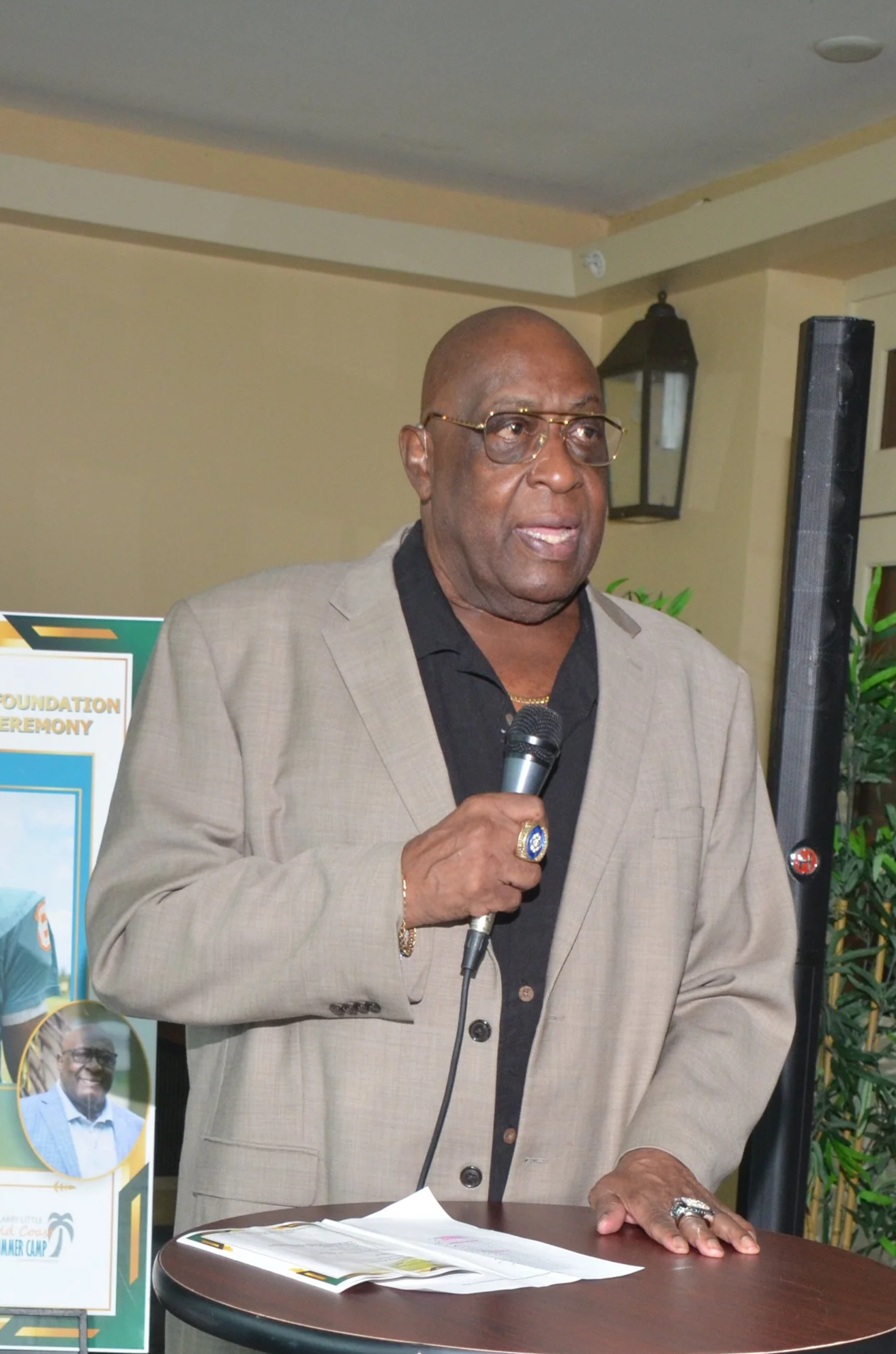 Man in a beige suit speaking at a podium during an awards ceremony with football-themed posters and a photo of a football game behind him.