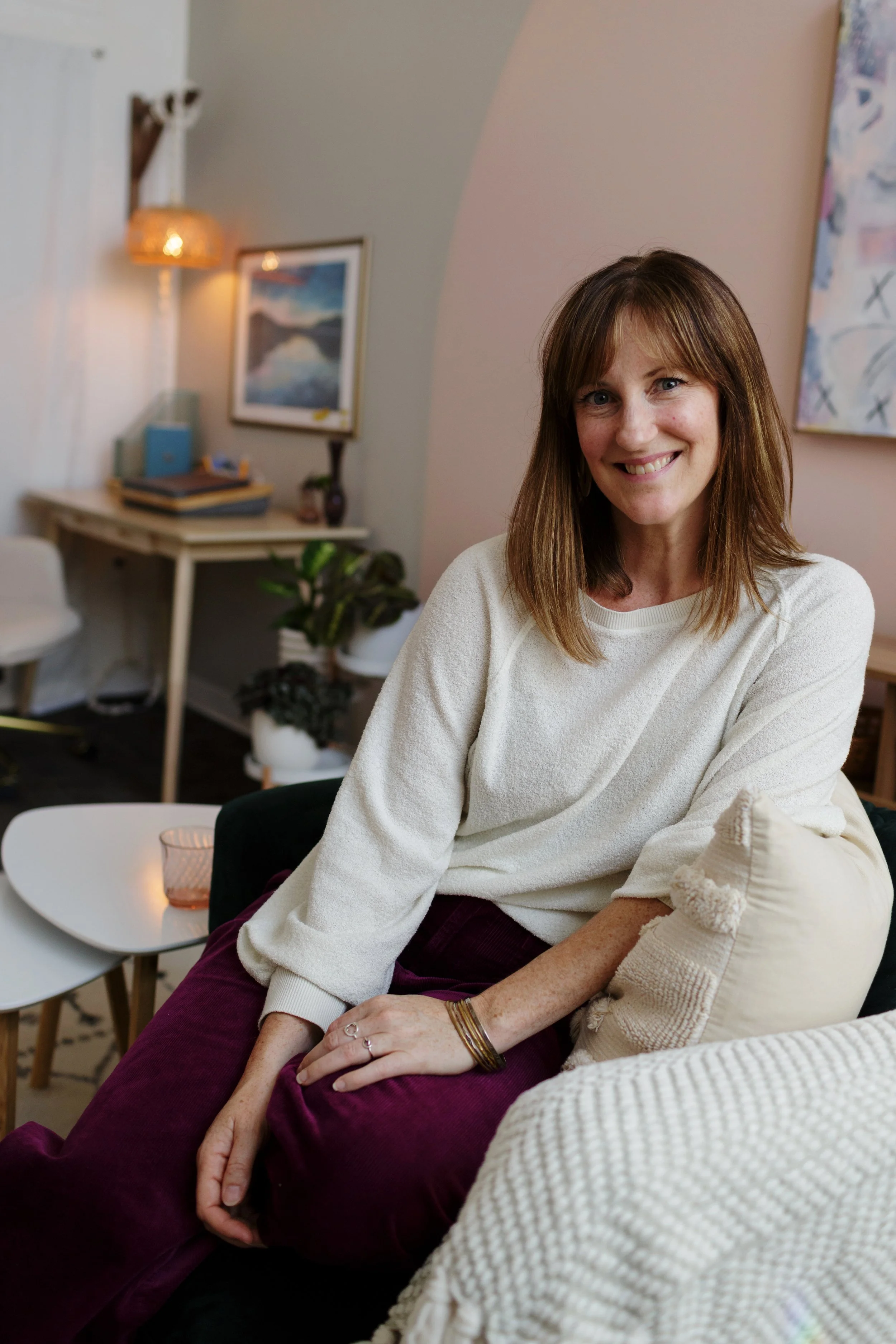 A woman with shoulder-length brown hair and a white sweater sitting on a beige sofa in a cozy living room, smiling.