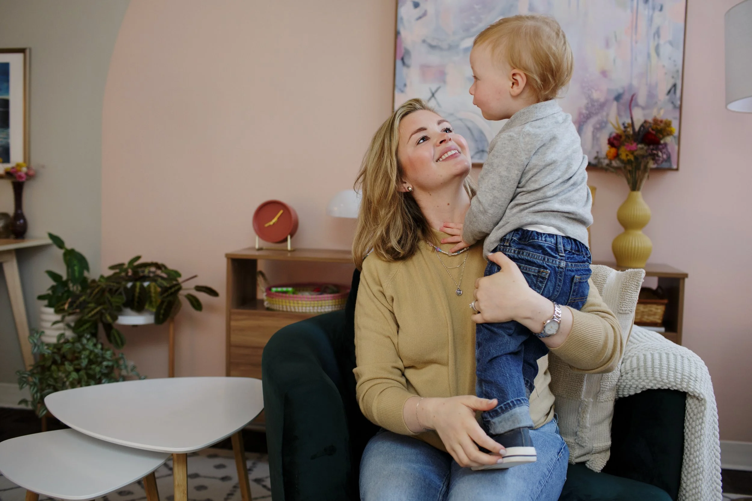 A woman with blonde hair and wearing a beige sweater is smiling and holding a young boy with blonde hair, wearing a gray sweater and jeans, while looking at each other in a living room.