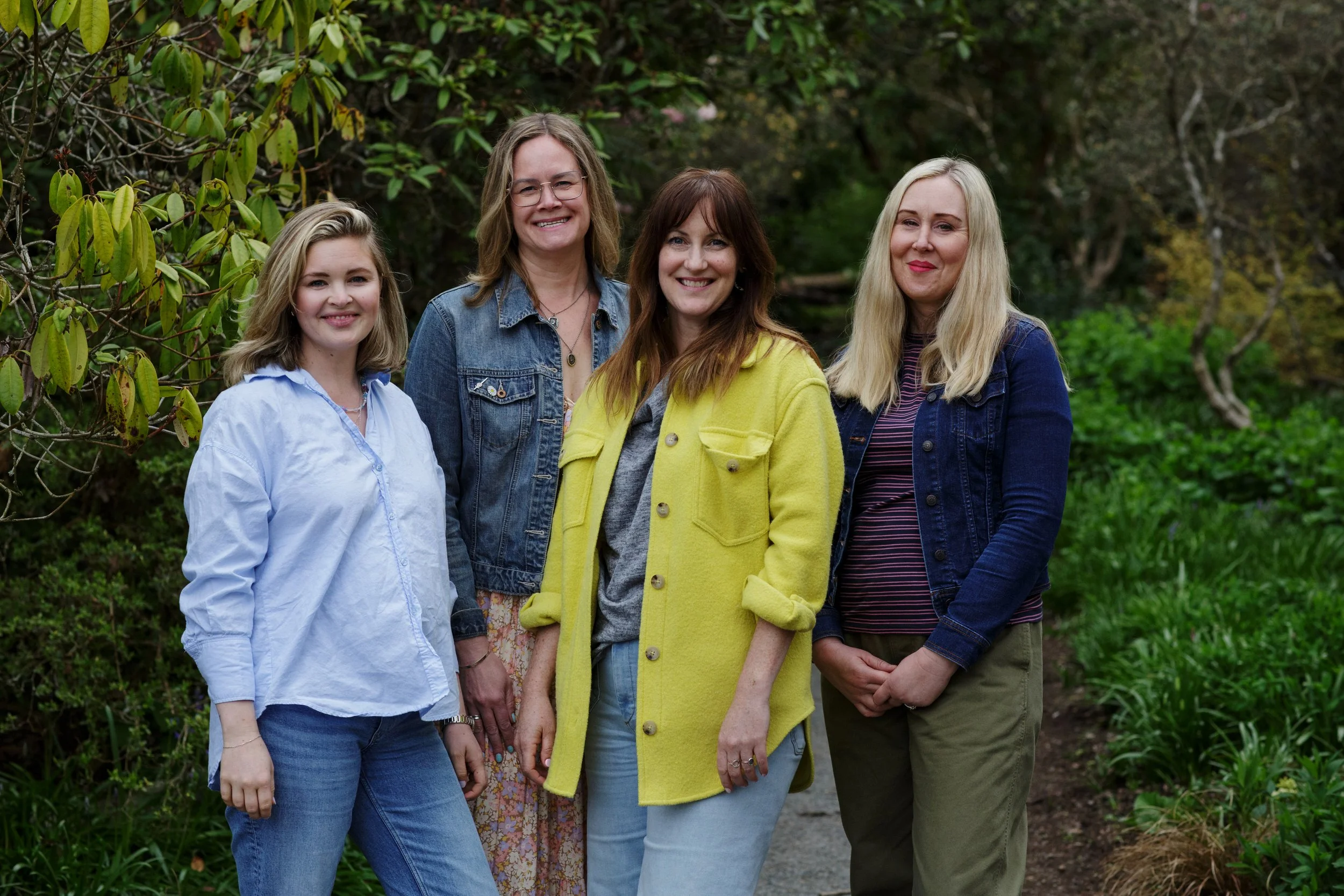 A group of five women smiling outdoors on a trail surrounded by greenery and trees.