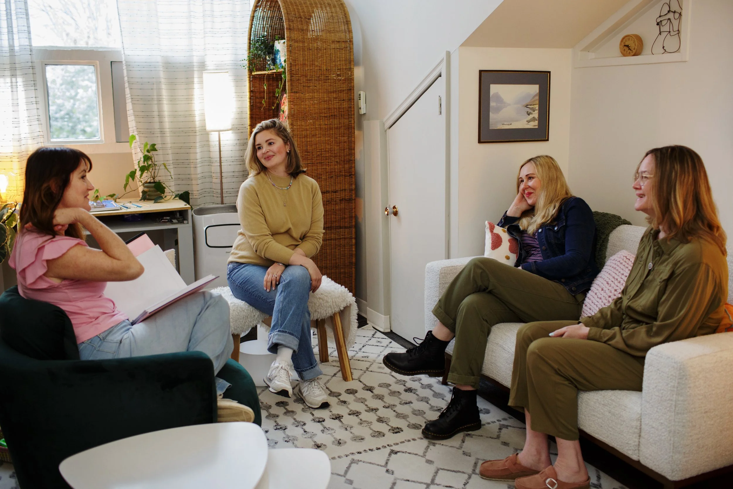 A group of five women sitting and engaging in conversation in a cozy living room with natural light, curtains, framed artwork, and indoor plants.