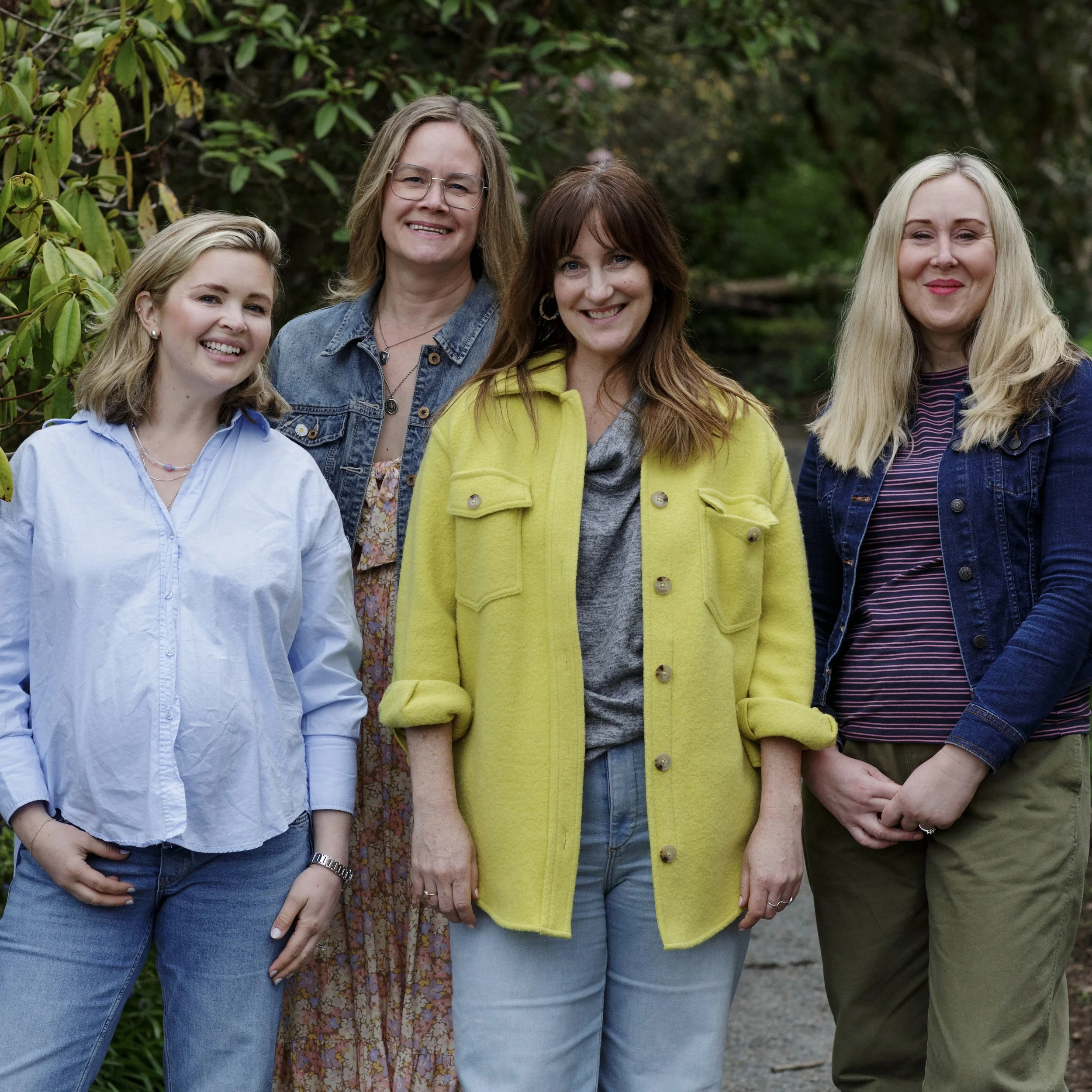 A group of five smiling women standing outdoors with greenery in the background.