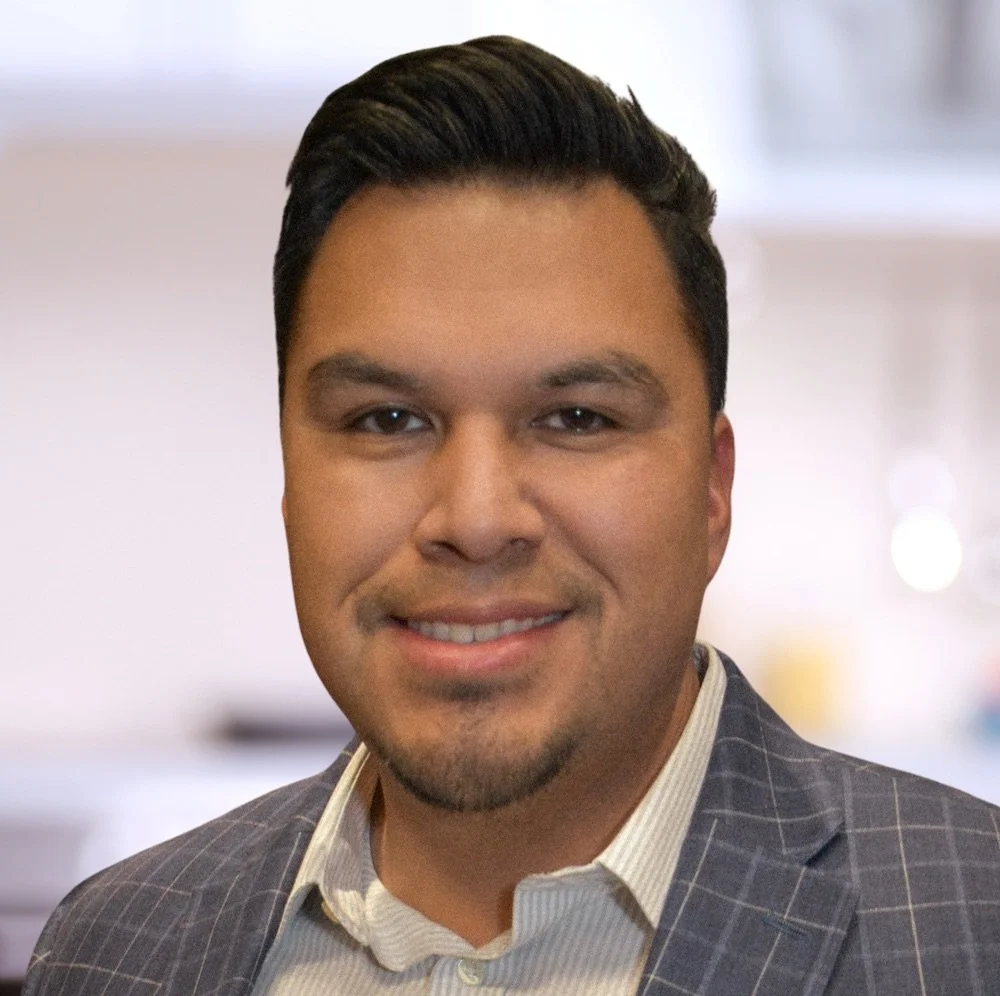 A man with dark hair, wearing a gray plaid suit jacket and a light-colored shirt, smiling for a photo in an indoor setting.