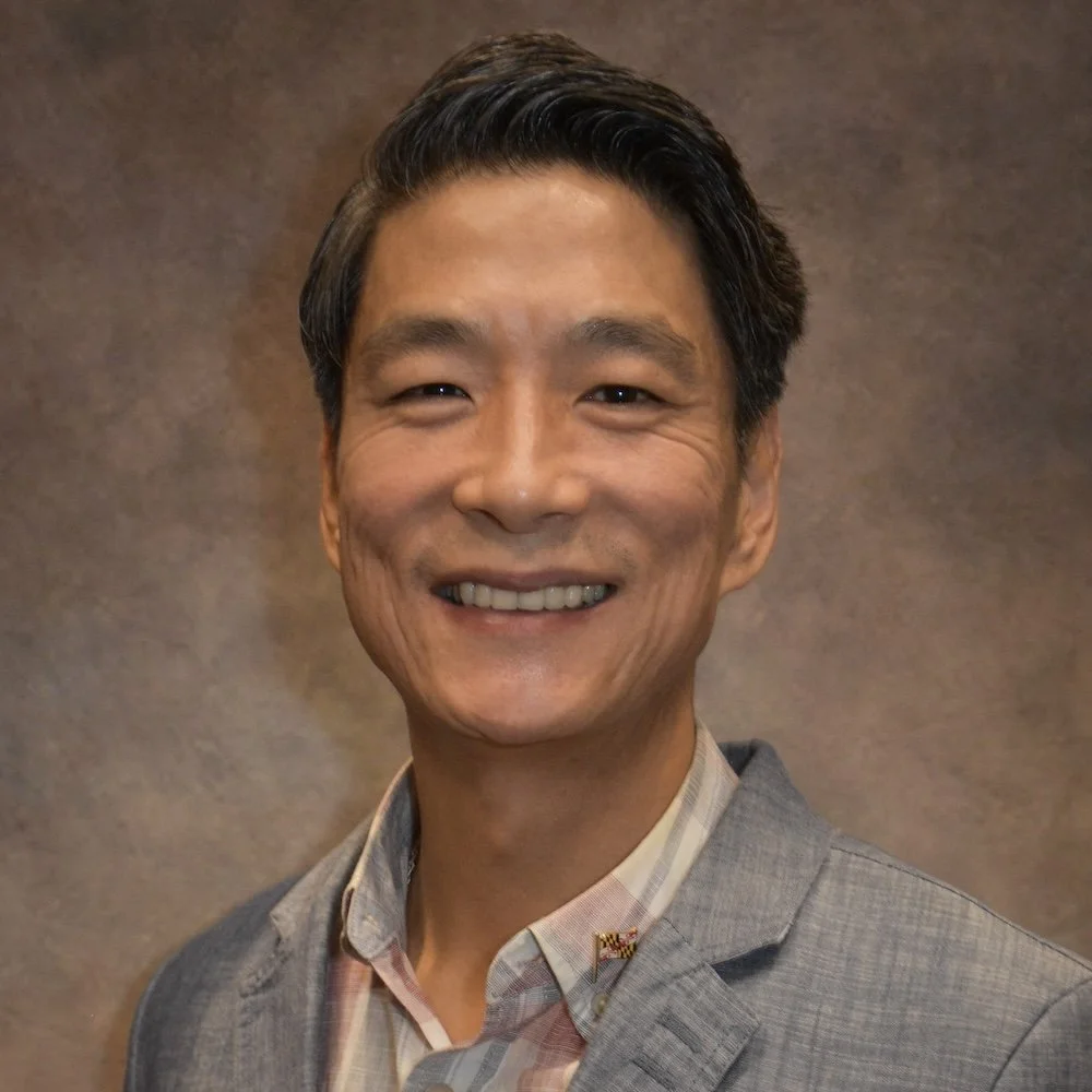 A smiling Asian man with short, dark hair wearing a grey suit jacket and a light-colored, checkered dress shirt against a neutral background.