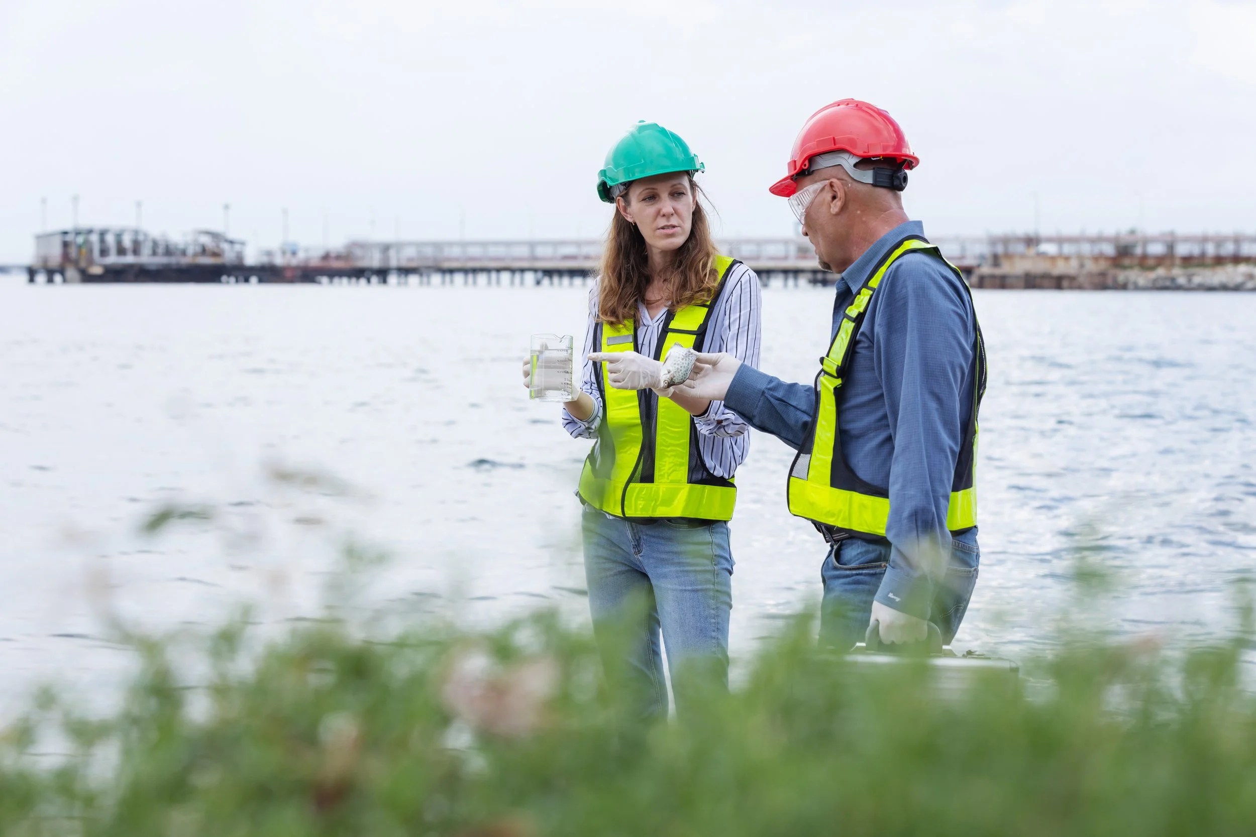 Two scientists, a woman and a man, wearing helmets and safety vests, are standing near water on a cloudy day. The woman is holding a glass container, and the man is pointing to a sample in his hand. They are discussing environmental or water analysis research.