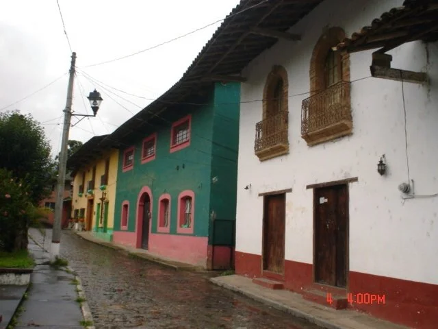 Colorful buildings on a cobblestone street in a small town, with vintage street lamps and trees, under an overcast sky.