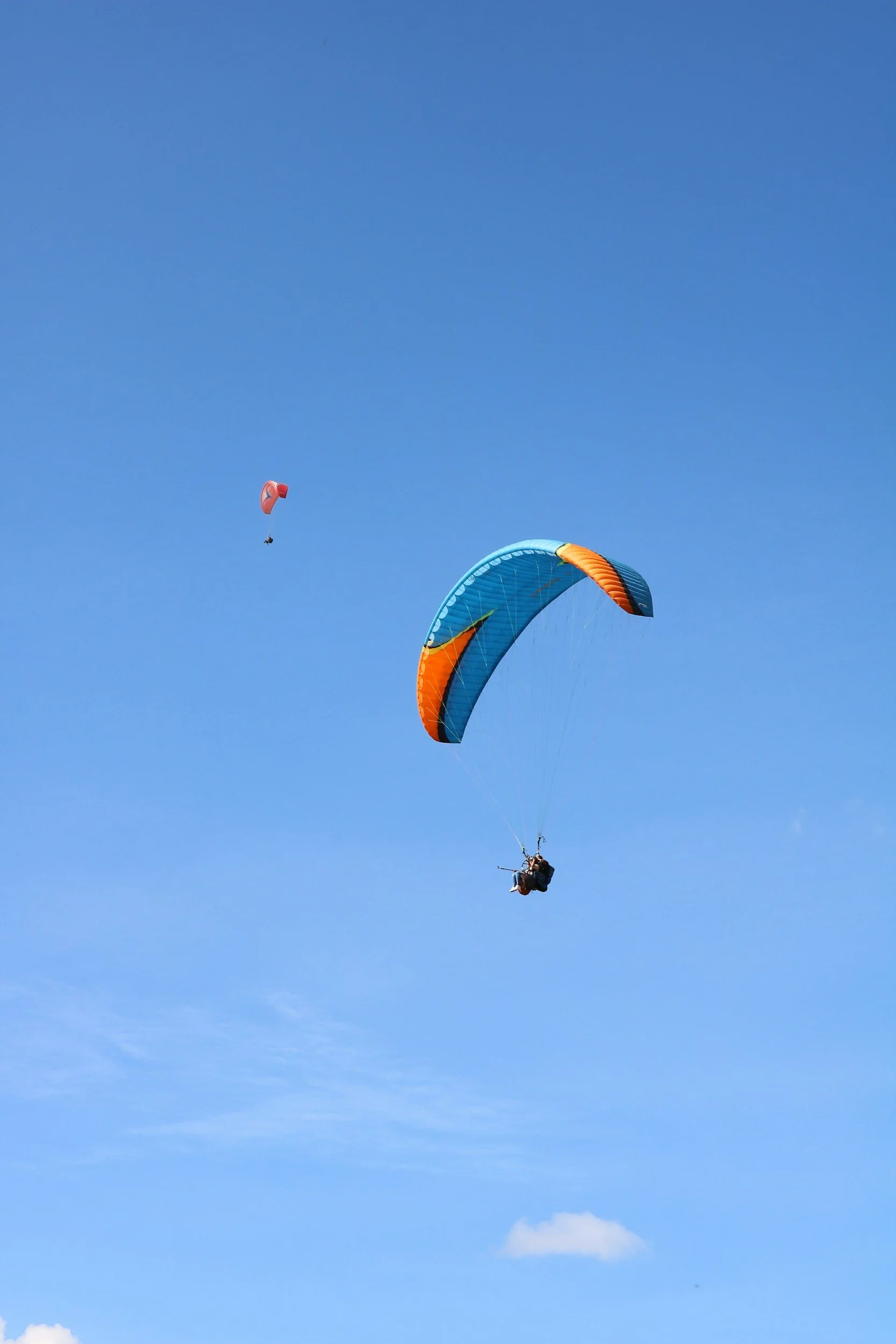 A person paragliding in the sky with a blue and orange parachute, and two other parachutes visible in the distance.