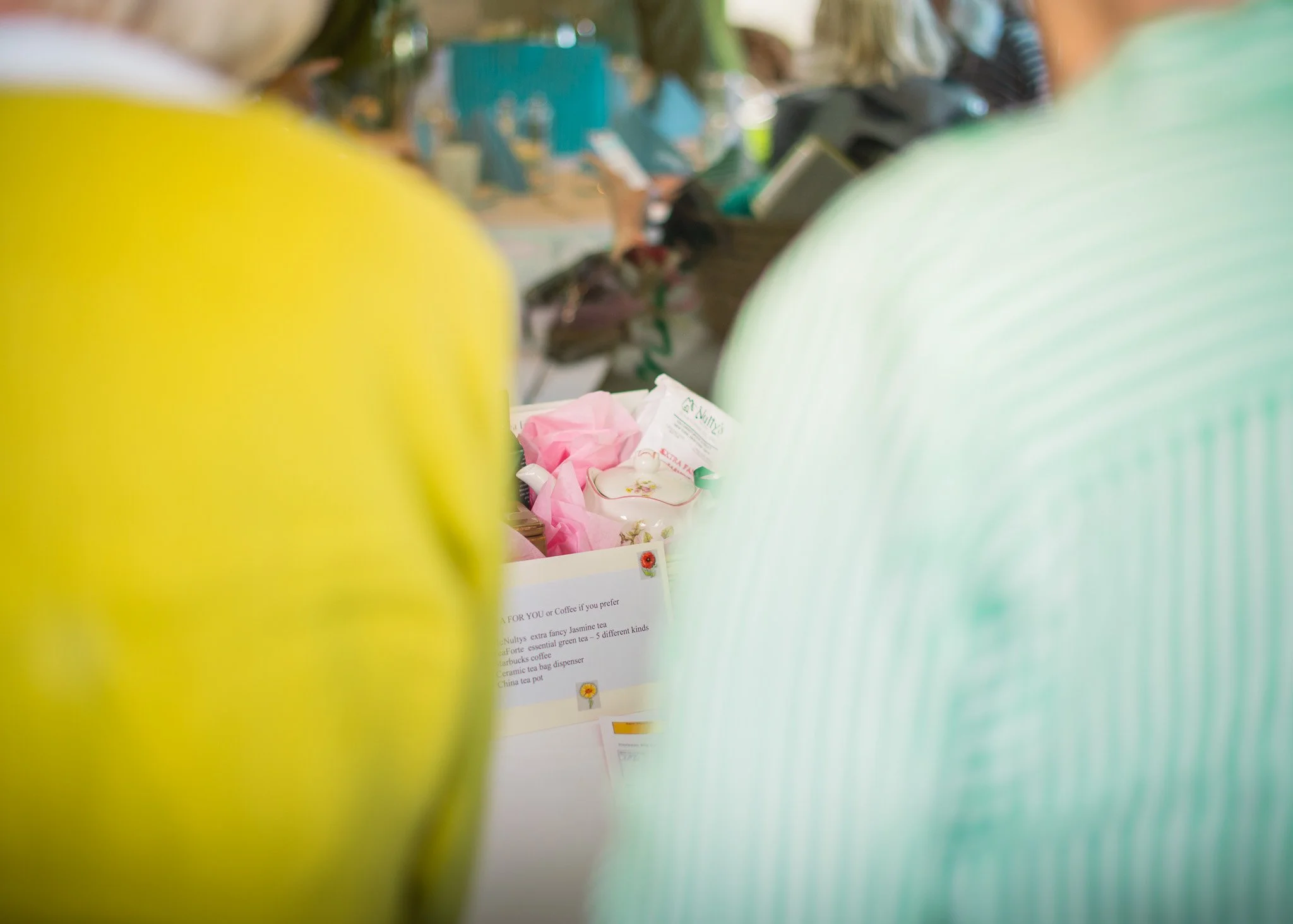 Blurred view of a display table at a store or cafe with pink tissue paper, a ceramic teapot, and a printed menu with tea options visible. The scene is viewed through yellow and green objects in the foreground.