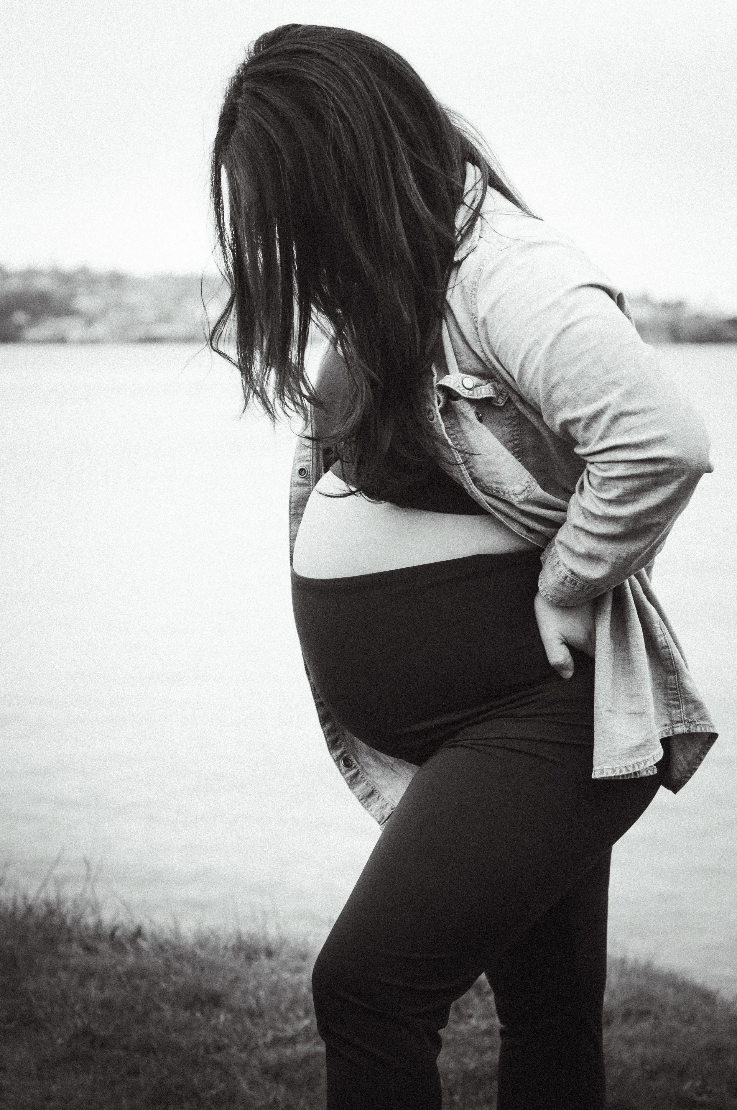 A pregnant woman with long dark hair, wearing a denim jacket and a black dress, stands outdoors near a body of water and looks down at her belly.
