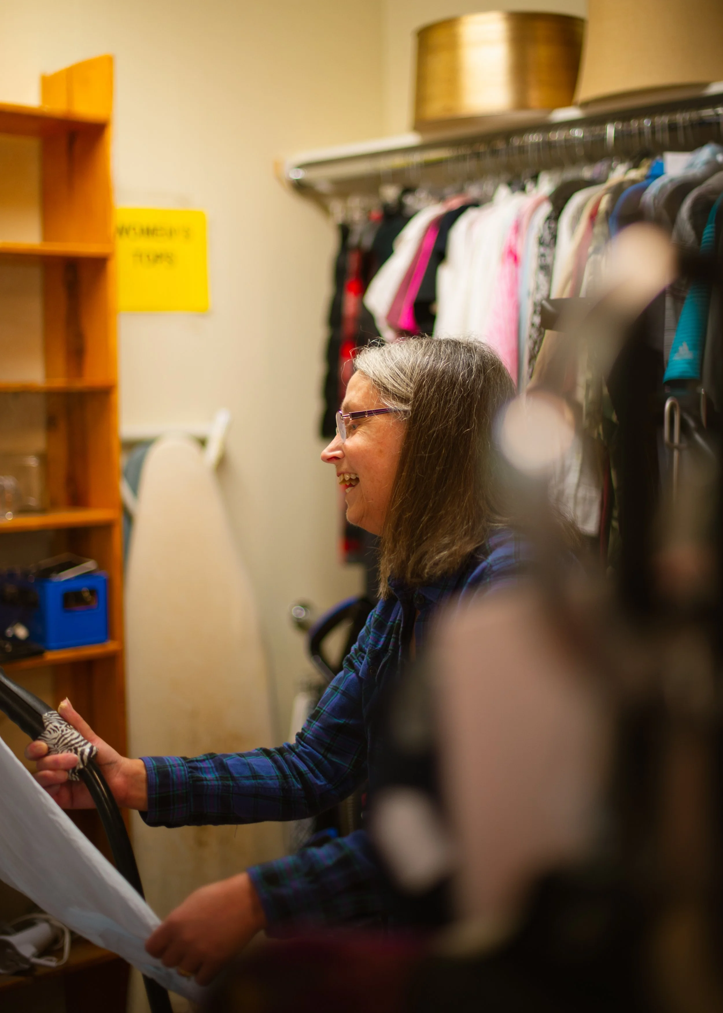 A woman with glasses and long hair, wearing a plaid shirt, smiling and looking at an electronic device in her hands inside a room with clothes on a rack and a yellow sign on the wall. in Seattle, Washington