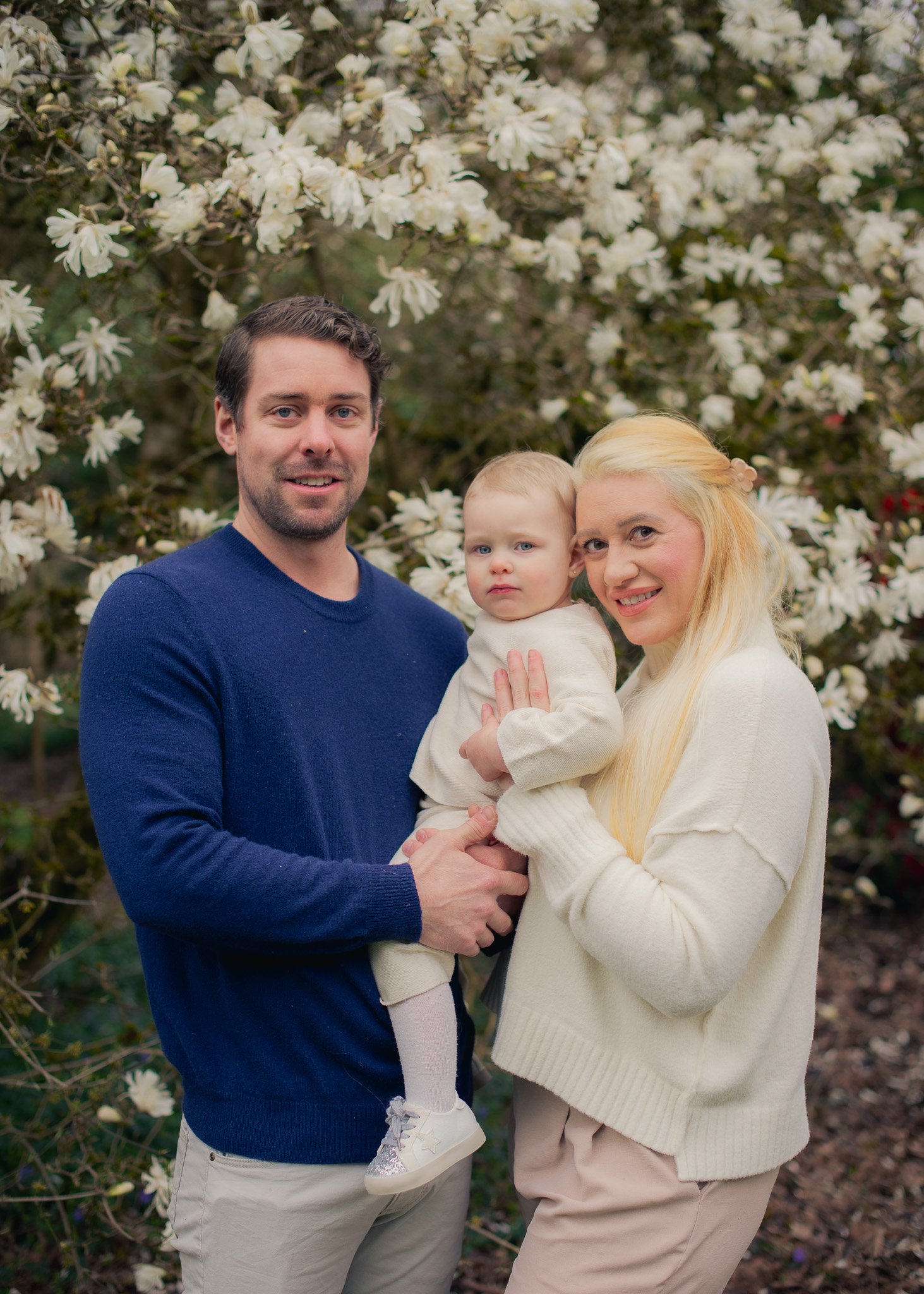 A family of three, a man, a woman, and a child, standing outside in front of blooming white flowers. The man has dark hair and a beard and is wearing a blue sweater. The woman has long blonde hair and is wearing a cream-colored sweater. The child has