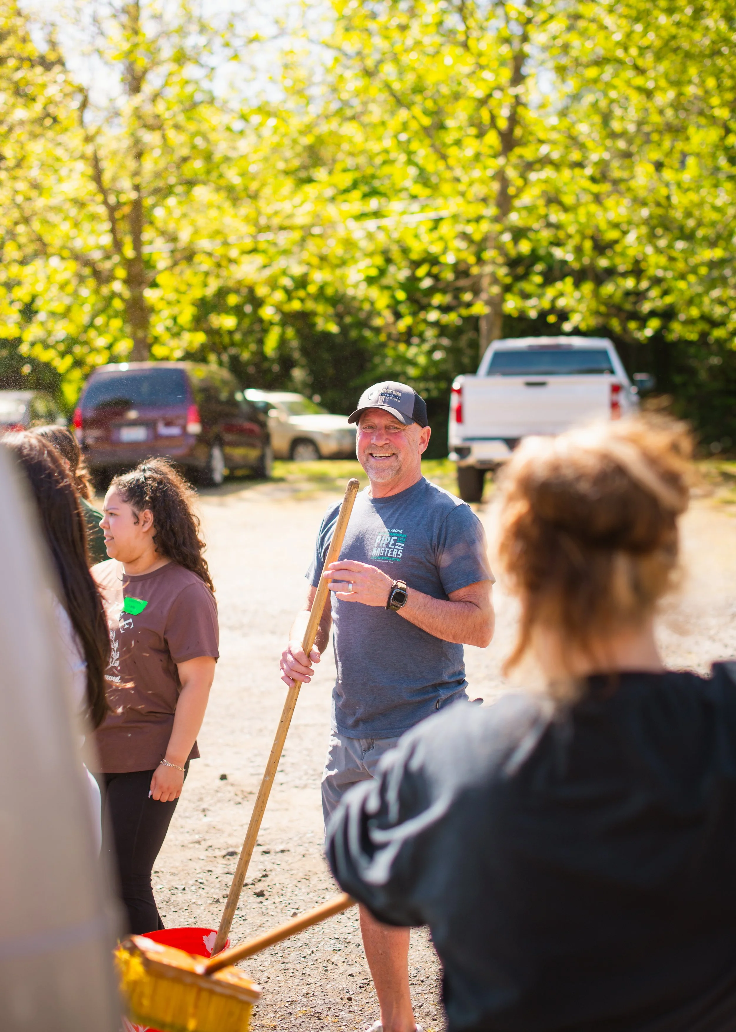 A group of people gathered outdoors on a sunny day, with a man smiling and holding a wooden stick, surrounded by others, in front of trees and parked vehicles. in Seattle, Washington