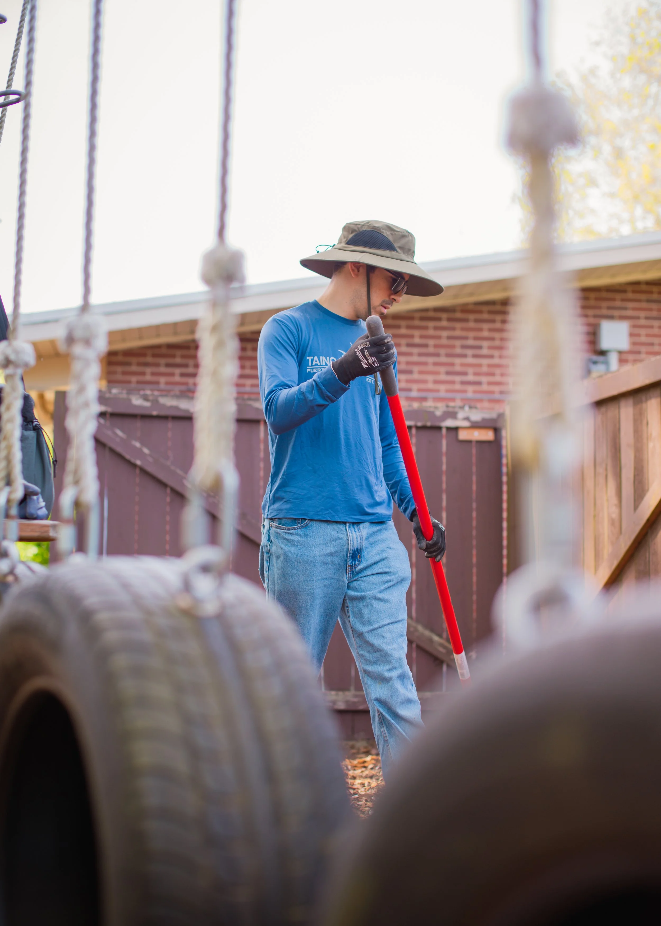 A man wearing a wide-brimmed hat, sunglasses, and gloves is holding a rake while working outdoors near a brick wall and wooden fence with ropes or chains hanging in the foreground. in Seattle, Washington 