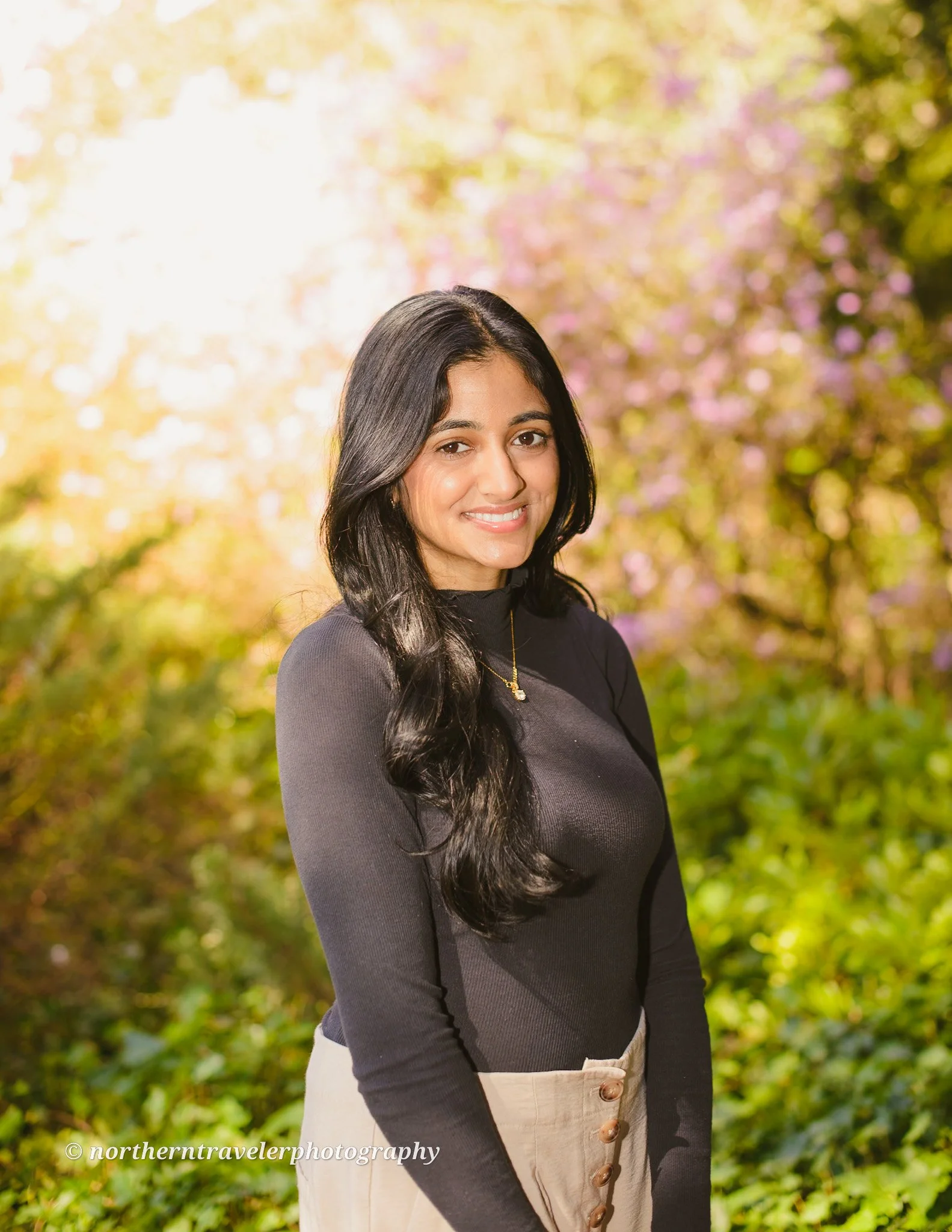 A woman with long dark hair smiling outdoors with trees and pink flowers in the background.