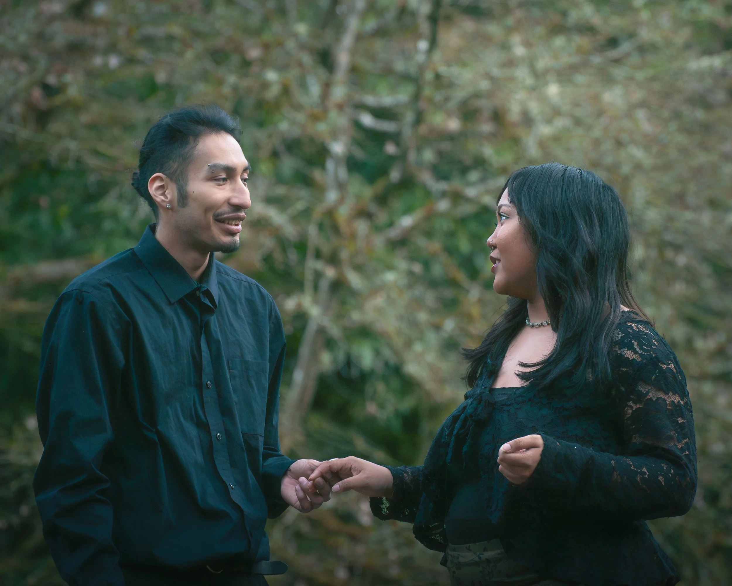 A man with dark hair and a woman with black hair are outdoors in a wooded area, holding hands and smiling at each other.
