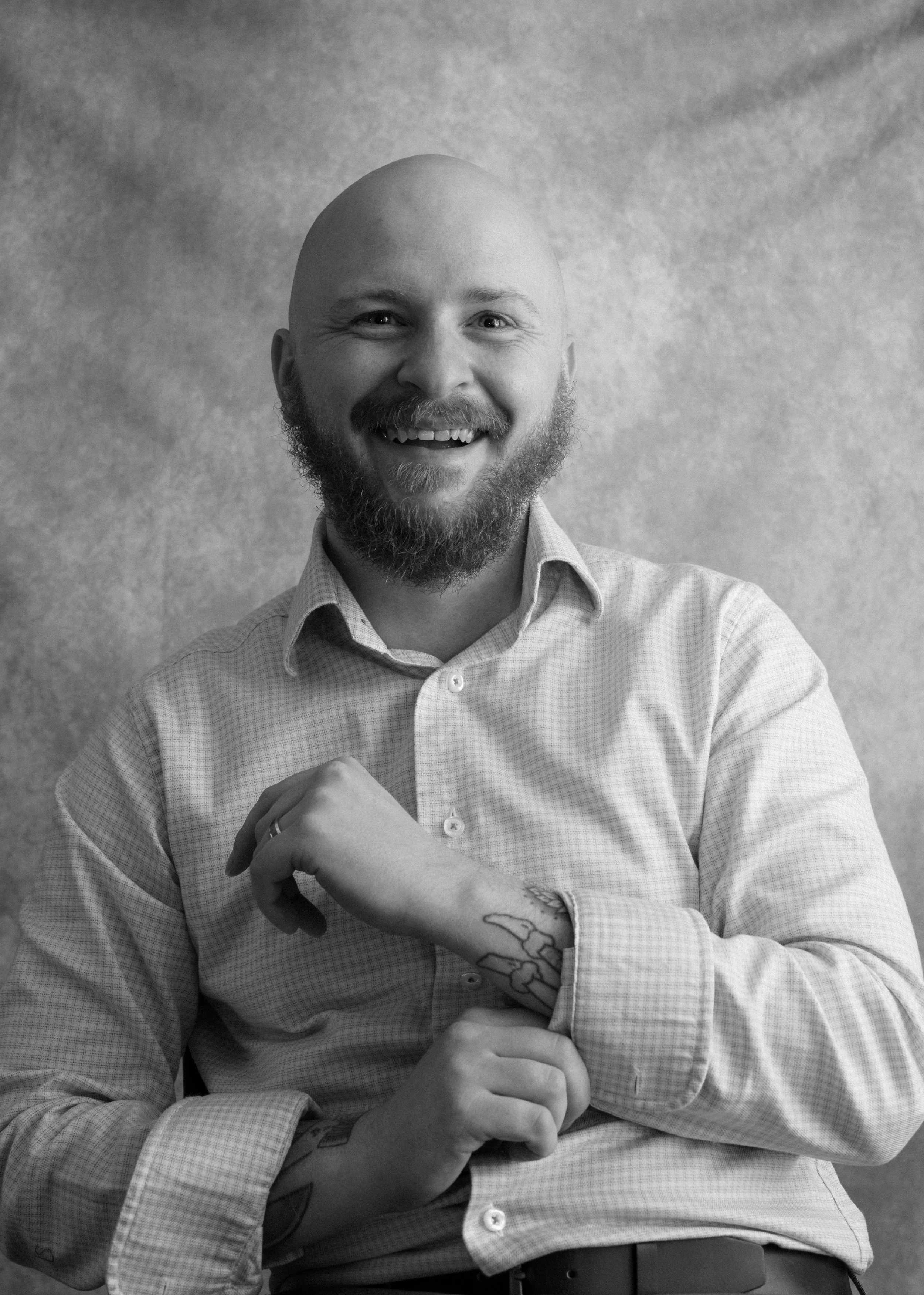 Black and white portrait of a smiling man with a beard, bald head, wearing a checkered shirt, making a fist with one hand showing a tattoo on his wrist.