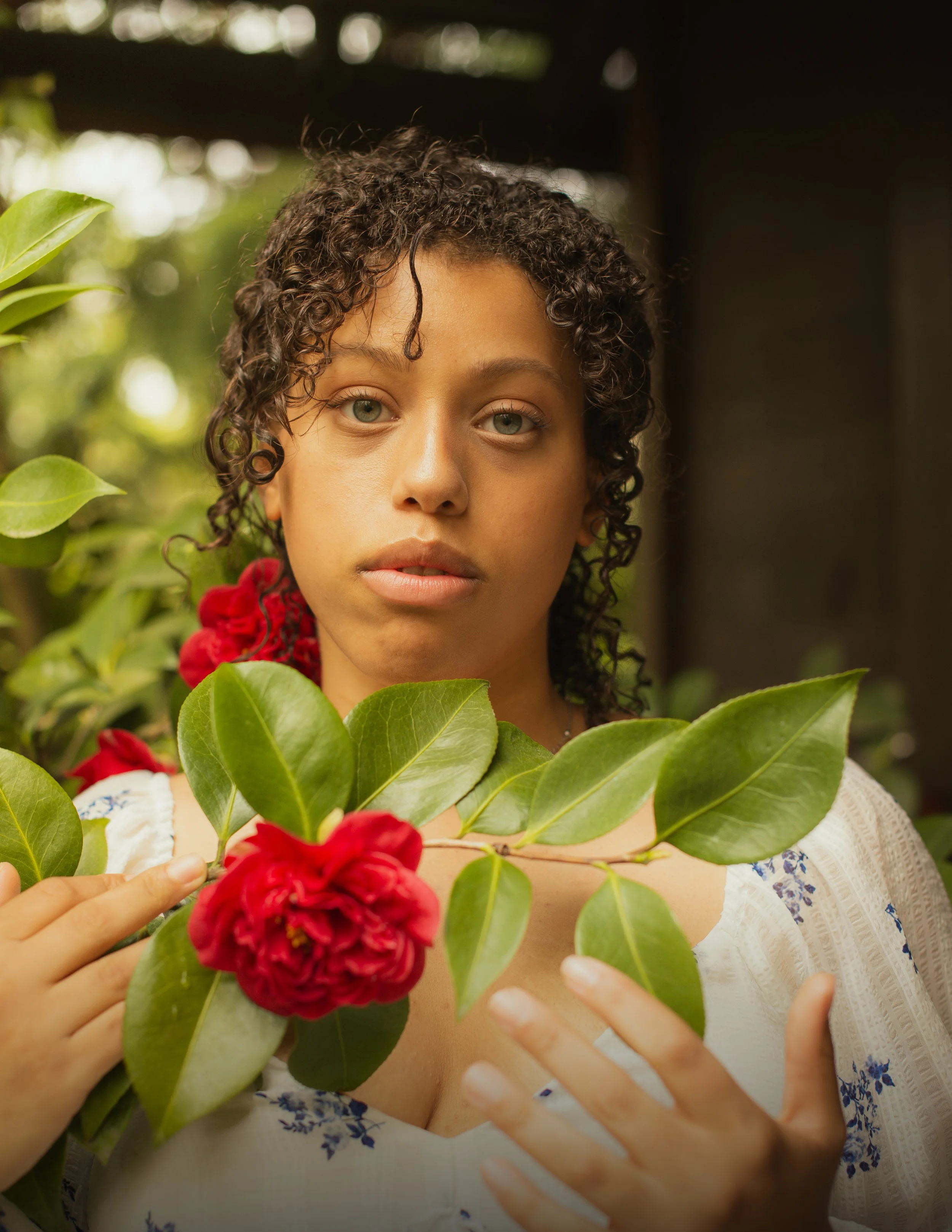 A woman with curly hair and green eyes holding a red flower and green leaves, wearing a white dress with blue floral patterns, and standing in front of a blurred green and black background.