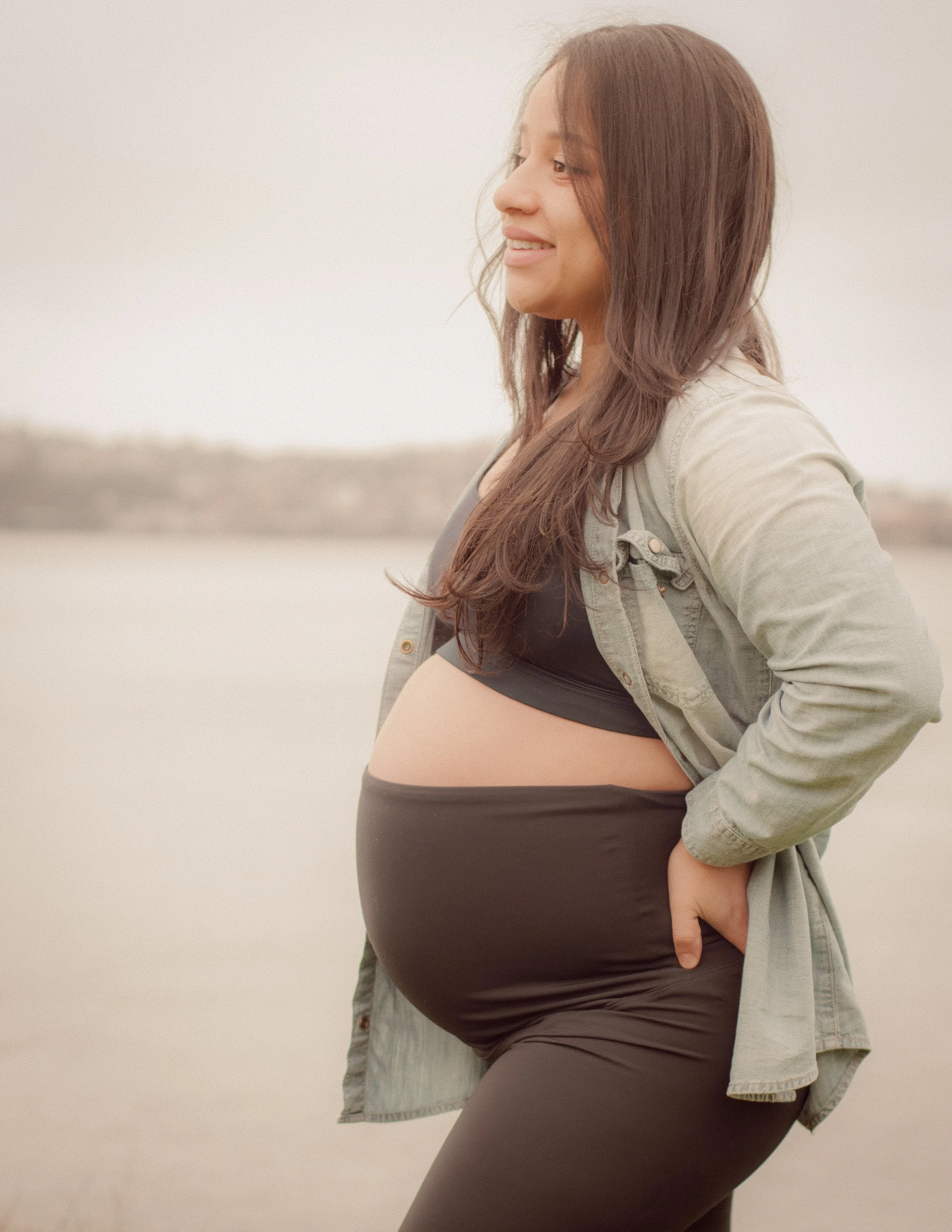 Pregnant woman standing outdoors, smiling, wearing a black sports bra, gray jacket, and black leggings, with a blurred natural background.