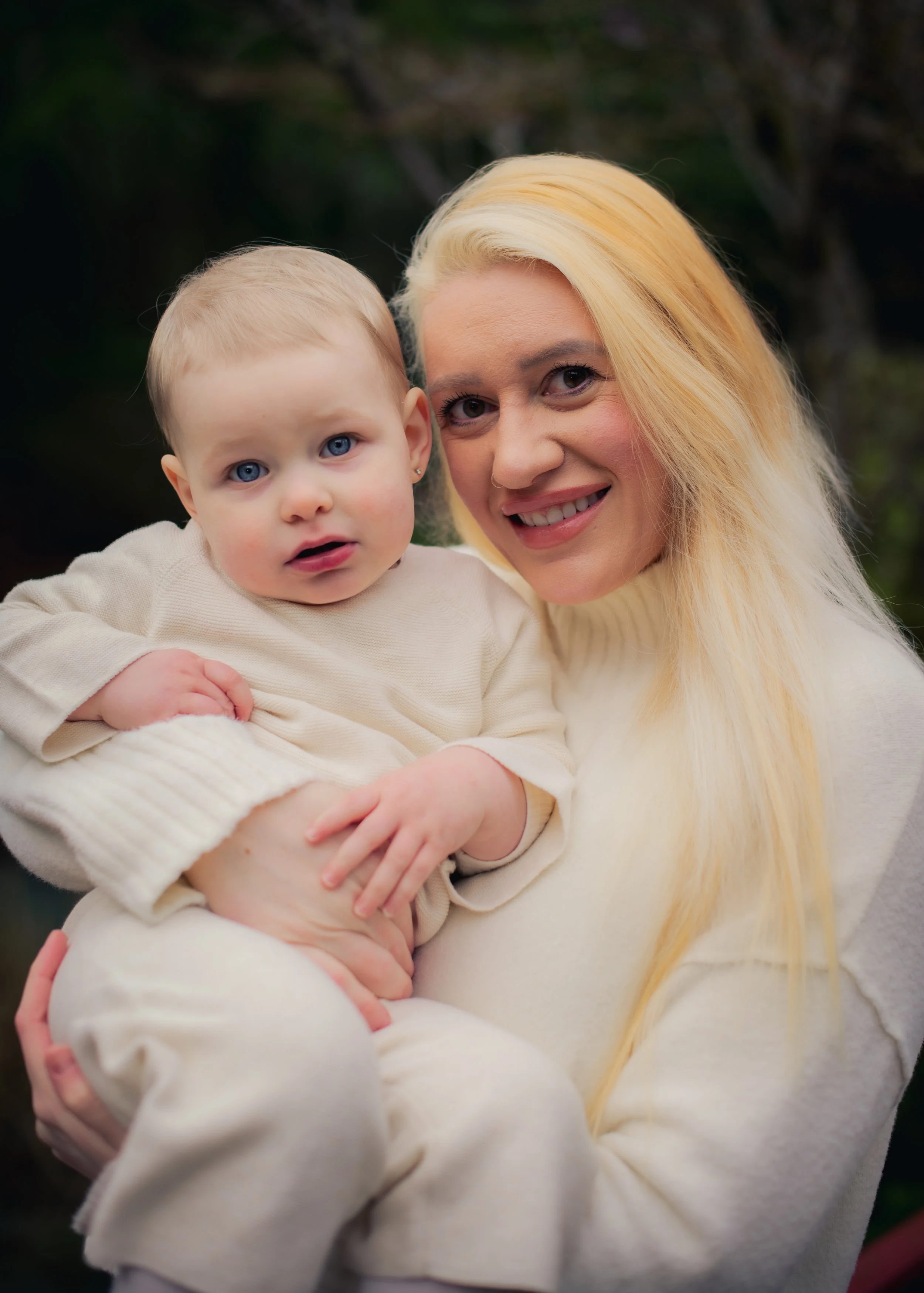 A woman with long blonde hair smiling and holding a young child with blonde hair and blue eyes outdoors.