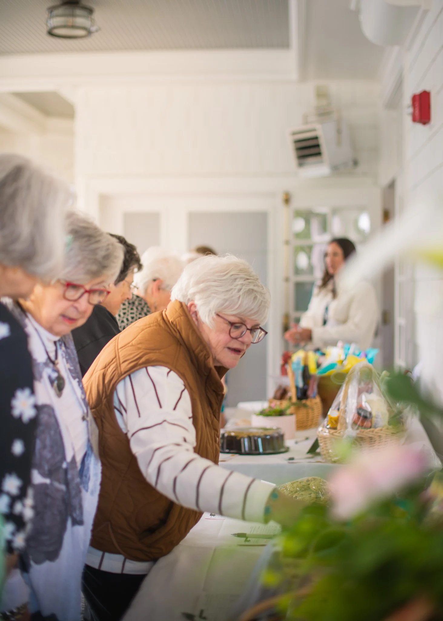 A group of older women at a table, examining plants or flowers, with a young woman in the background behind a display of various plants and items.