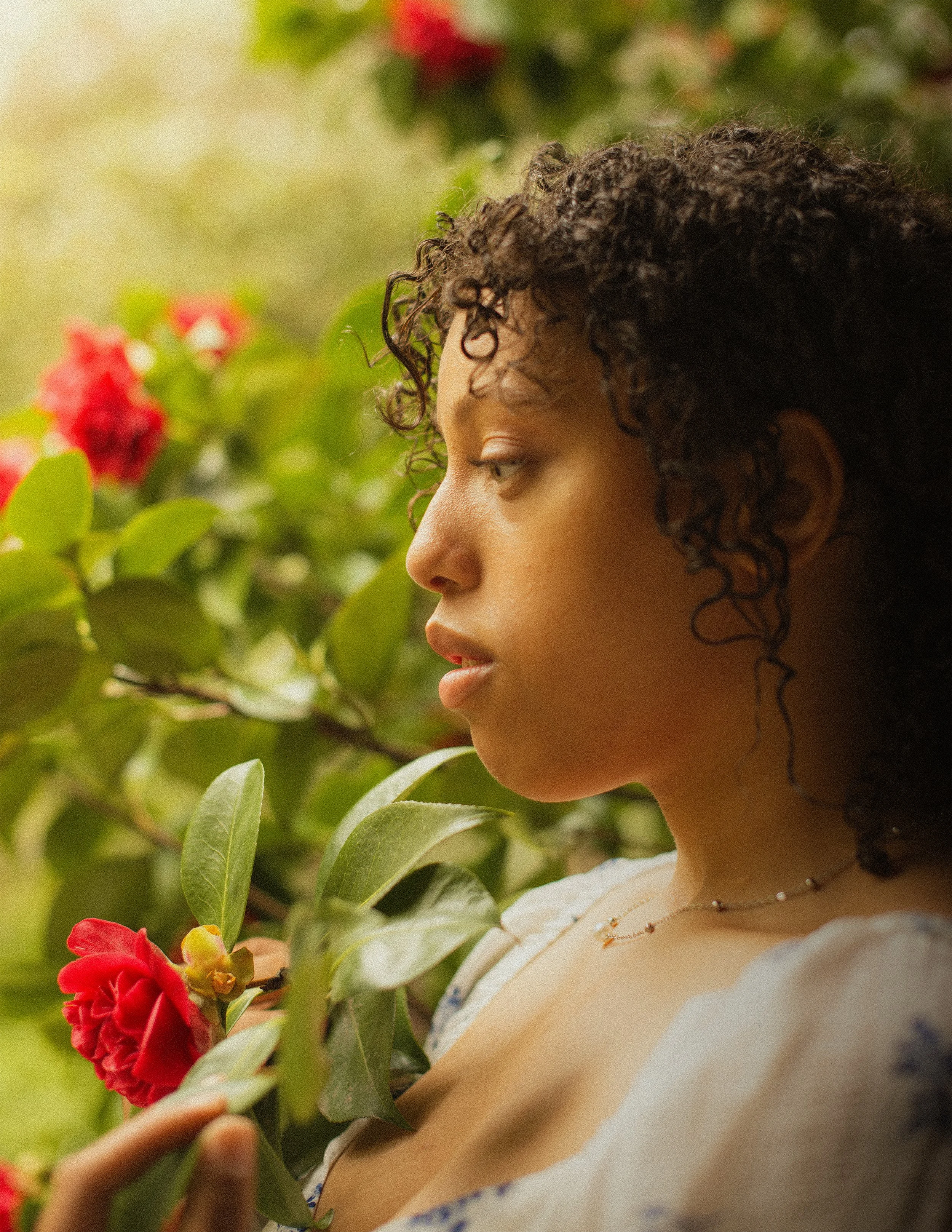 A woman with curly hair is holding a red flower in a lush garden.