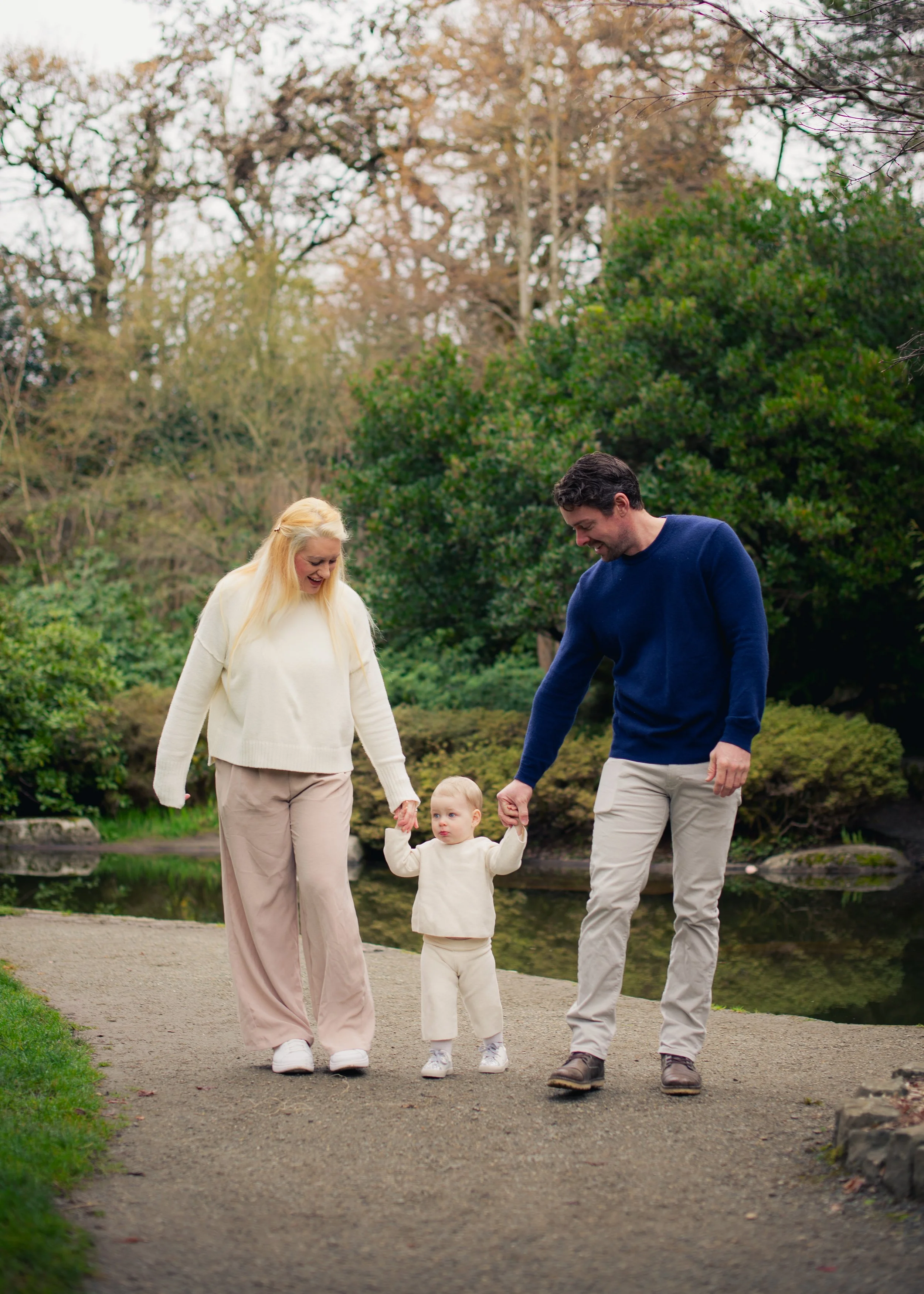 A family of three walking hand in hand along a park path near a pond, with trees and greenery in the background.